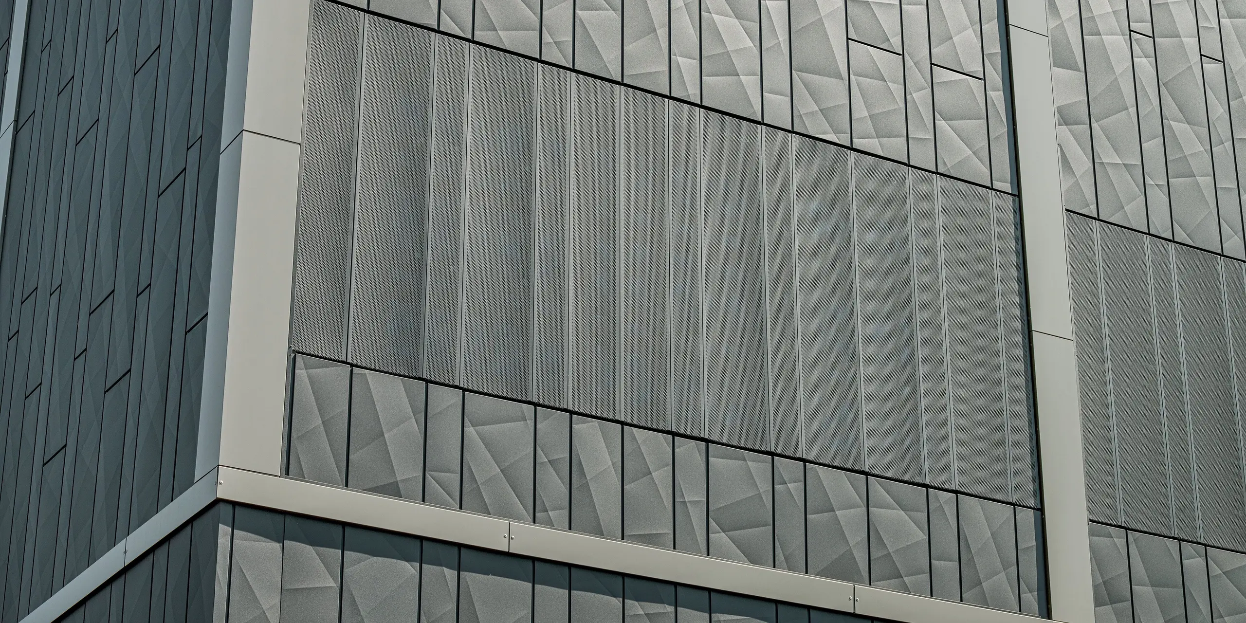 L'image présente une coupe détaillée de la façade en aluminium du centre commercial de Rohrbach. Les panneaux Siding.X anthracite sont particulièrement remarquables : leur structure géométrique et leurs surfaces variées créent une impression de dynamisme et de modernité. Des éléments Siding perforés sont visibles dans la partie inférieure de l'image. Outre leur aspect esthétique, ils assurent une ventilation ciblée et une transmission optimale de la lumière. L'association de panneaux lisses et perforés confère à la façade un design à la fois fonctionnel et esthétique, alliant esthétique et praticité.