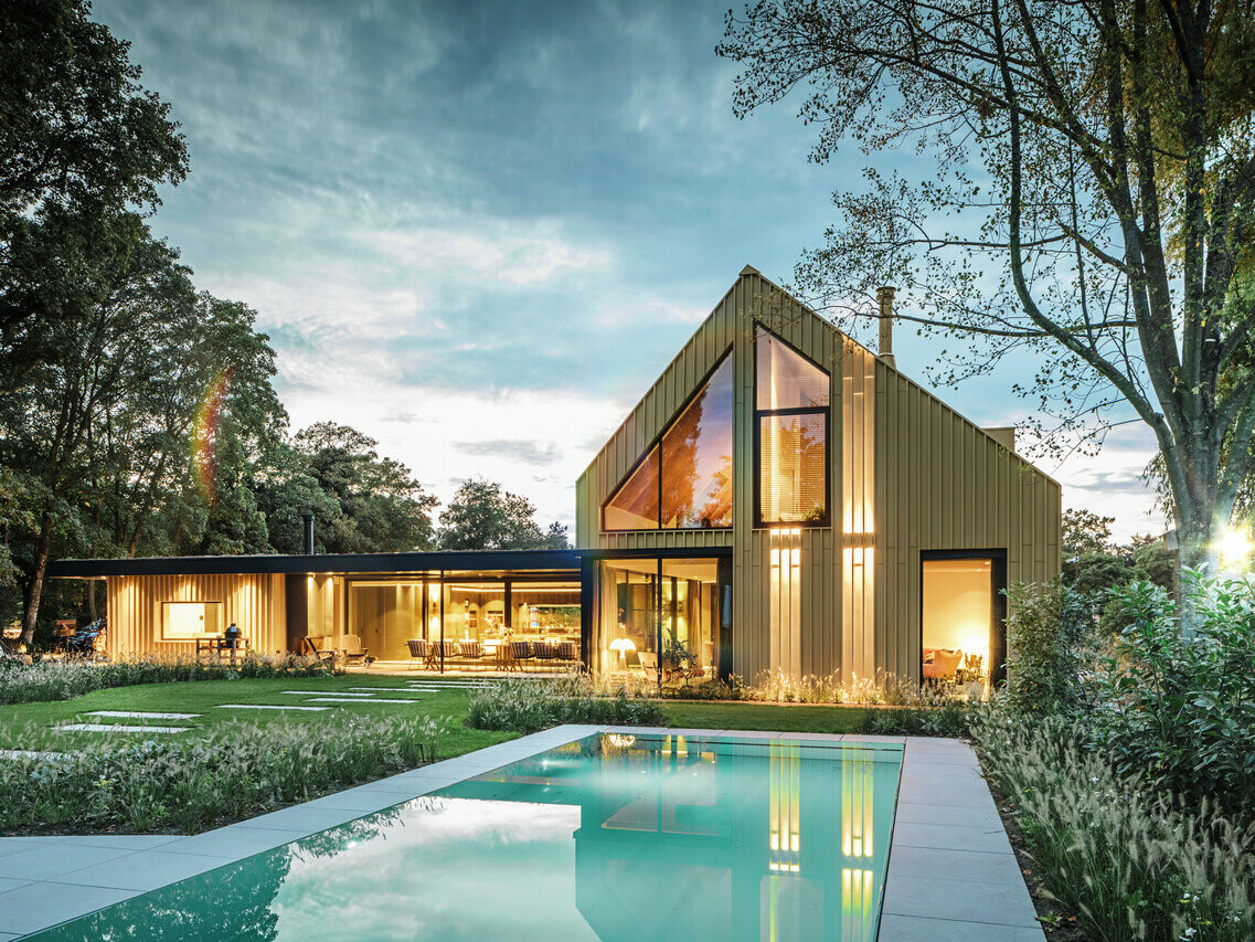 Vue de la maison individuelle d'Elst recouverte en Prefalz et Falzonal. La maison est entourée d'arbres et une piscine est visible au premier plan.