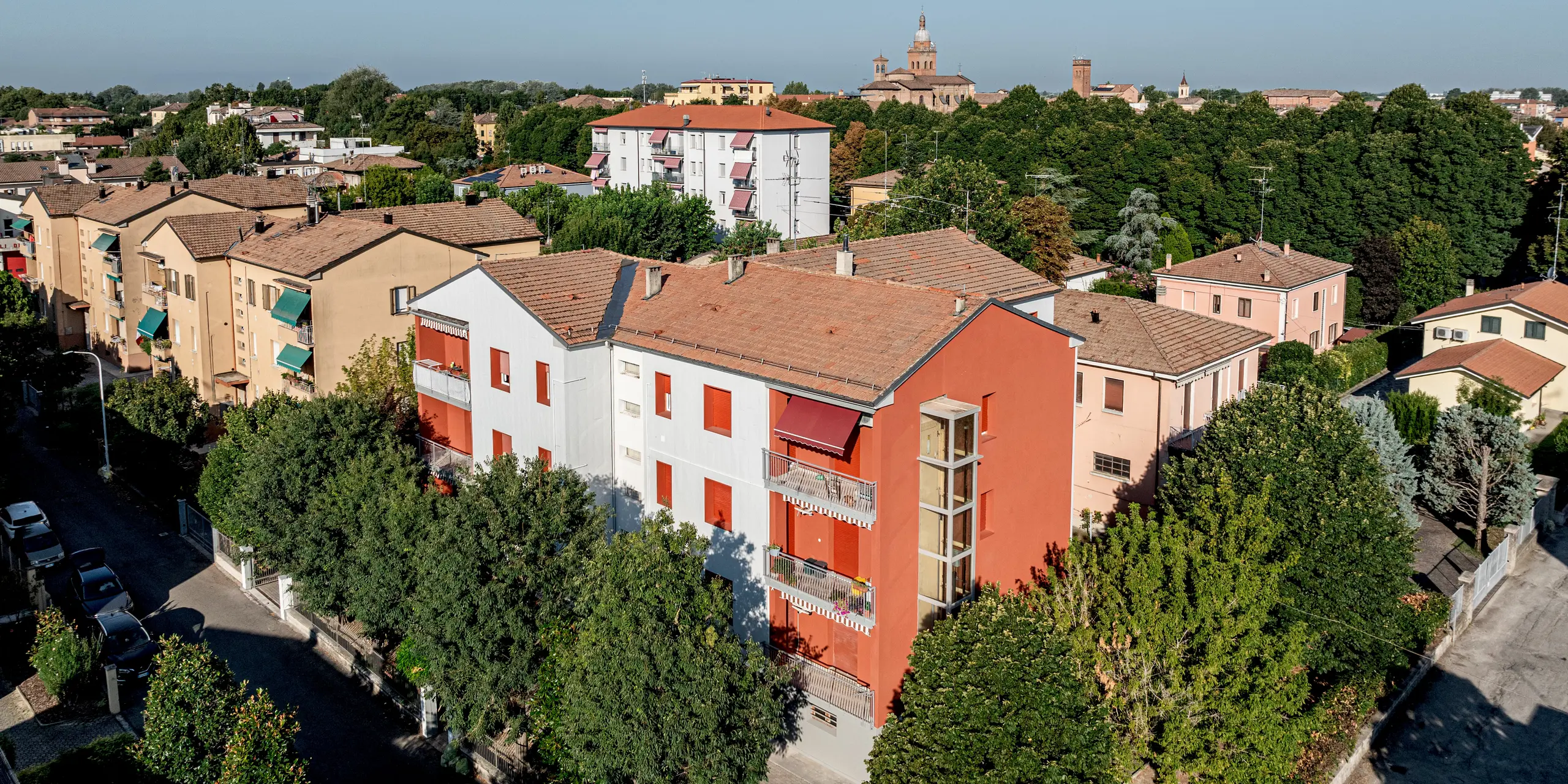 Vue aérienne du complexe résidentiel Alba B, situé dans un quartier résidentiel verdoyant bordé d'une dense rangée d'arbres et entouré de bâtiments aux toits de tuiles. Le bâtiment rénové se distingue nettement par sa façade lumineuse en aluminium PREFA, réalisée avec le système Siding.X. Les panneaux verticaux, disposés en panneaux, révèlent une texture fine et dynamique grâce au jeu d'ombres et de lumières, tandis que les encadrements de fenêtres et les balcons rouges apportent une touche d'élégance. La solution de façade PREFA séduit par sa grande résistance aux intempéries, sa durabilité et son entretien minimal, tout en conservant une esthétique moderne et épurée.