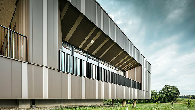Vue détaillée de la façade moderne en aluminium du centre événementiel ZIB Nikolai à St. Veit, avec des rayures verticales dans les tons marron et beige-gris. La loggia aux garde-corps noirs et aux fenêtres spacieuses offre une vue sur la verdure, tandis que la façade impressionne par ses lignes claires et ses formes géométriques. En arrière-plan, vous pouvez voir un paysage naturel sous un ciel légèrement nuageux.