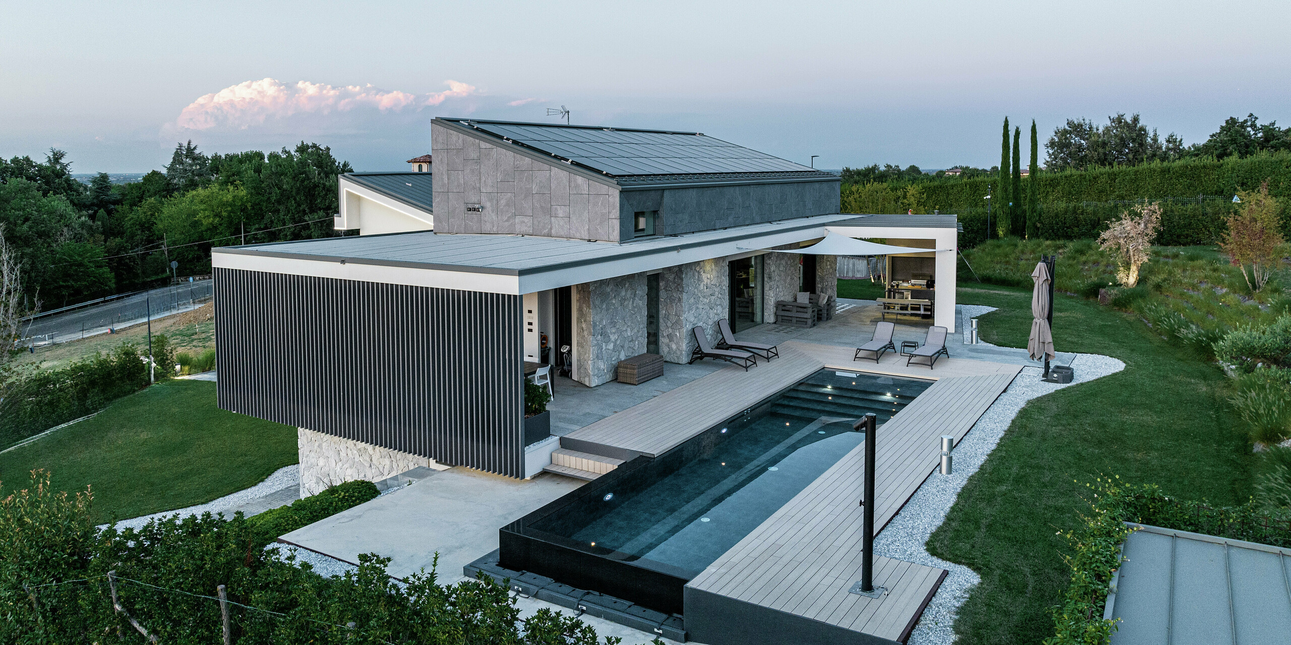 Vue latérale du luxueux immeuble résidentiel avec vue sur l'élégant toit PREFALZ en P.10 anthracite. La solution en aluminium durable de PREFA garantit une toiture durable et facile d'entretien avec un design moderne. La spacieuse terrasse couverte avec coin salon et piscine à débordement crée une atmosphère de bien-être exclusive en harmonie avec la nature environnante. L'installation photovoltaïque alimente la maison en électricité respectueuse de l'environnement grâce à l'énergie solaire.