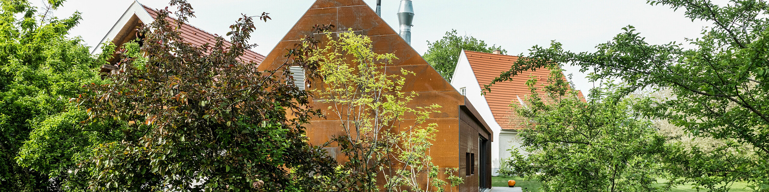 Résidence Velich dans le Seewinkel, Burgenland : Vue sur le jardin moderne agrémenté de belles plantes. La résidence sert de lieu pour le dialogue PREFARENZEN 2025, lors duquel architectes et experts PREFA sélectionnent les projets d’architecture les plus intéressants. Au premier plan, on observe un élégant mur métallique à la patine rouille, sur lequel est inscrit « RÉSIDENCE VELICH ». L'architecture du bâtiment marie habilement tradition et modernité, s’intégrant harmonieusement dans le paysage naturel.