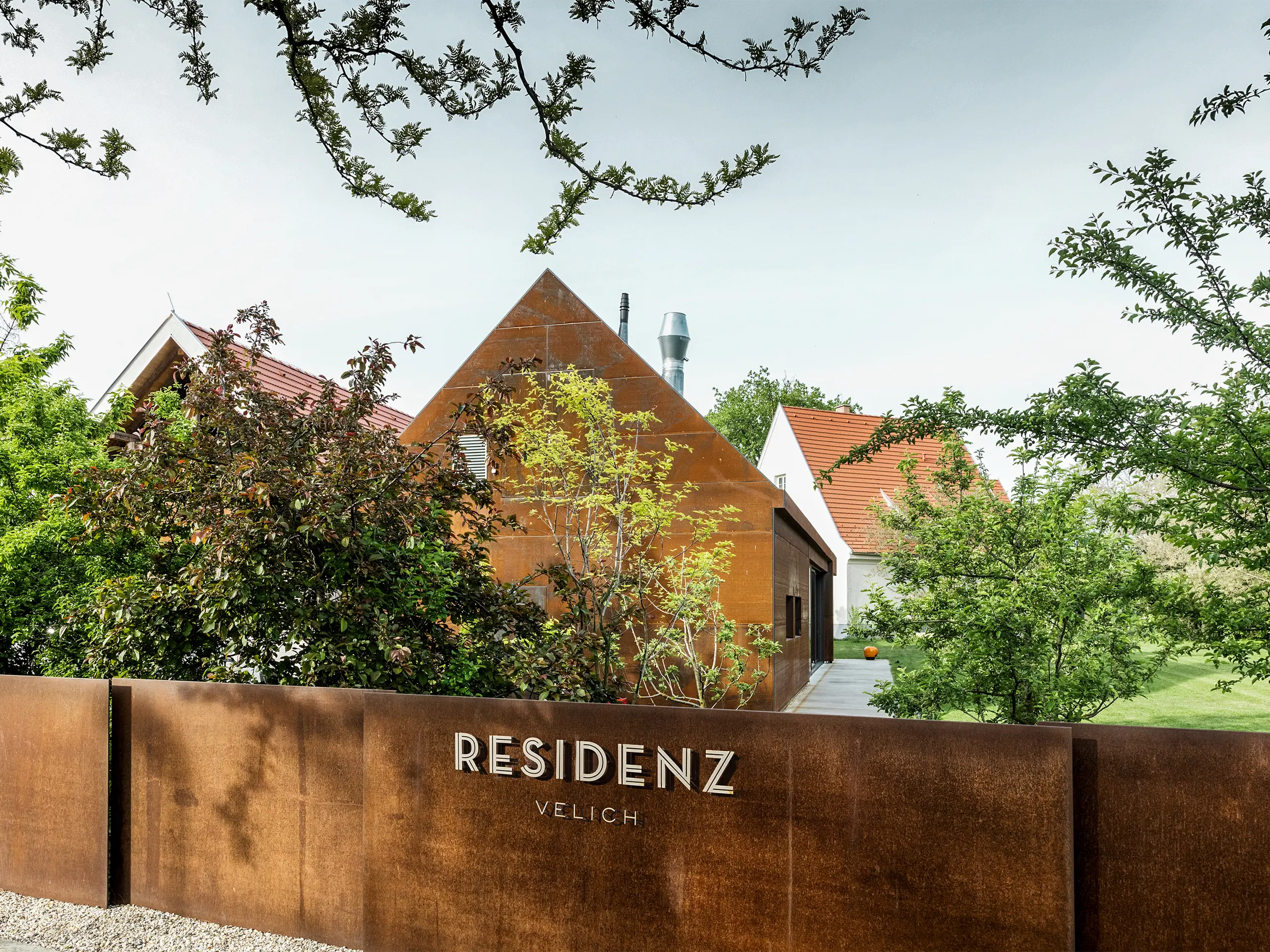 Résidence Velich dans le Seewinkel, Burgenland : Vue sur le jardin moderne agrémenté de belles plantes. La résidence sert de lieu pour le dialogue PREFARENZEN 2025, lors duquel architectes et experts PREFA sélectionnent les projets d’architecture les plus intéressants. Au premier plan, on observe un élégant mur métallique à la patine rouille, sur lequel est inscrit « RÉSIDENCE VELICH ». L'architecture du bâtiment marie habilement tradition et modernité, s’intégrant harmonieusement dans le paysage naturel.