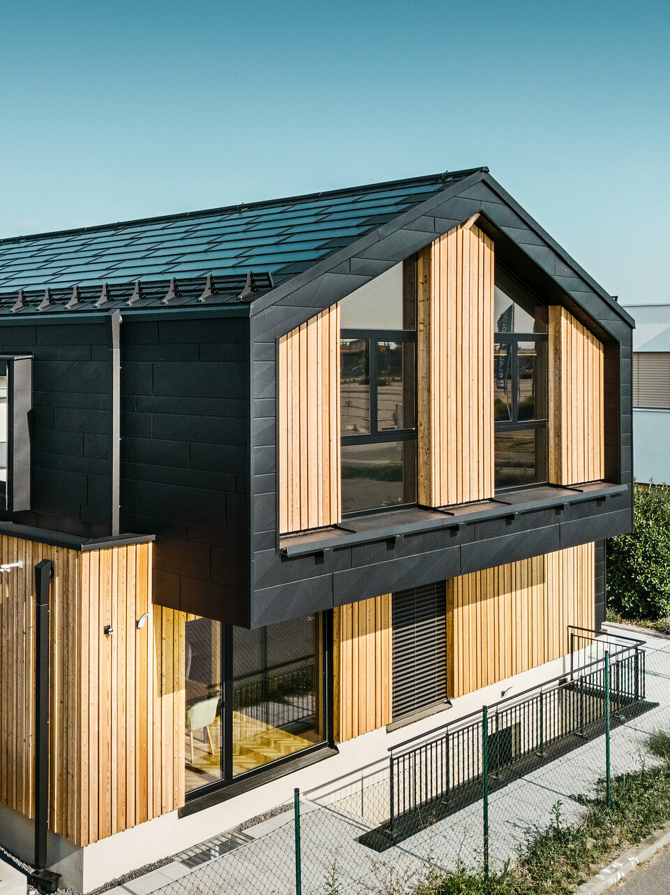 Façade de la maison modèle Autark dans le centre de maisons préfabriquées Blaue Lagune, Vösendorf (AT). Le toit est recouvert de tuiles solaires PREFA noirs et de R.16 avec un revêtement P.10 de haute qualité. La façade est constituée d'une combinaison d'éléments verticaux en bois et de PREFA Siding.X en P.10 noir, qui confère au bâtiment un caractère moderne et durable. Le drapeau de la société Erler Bau GmbH flotte sur le bord droit de l'image.