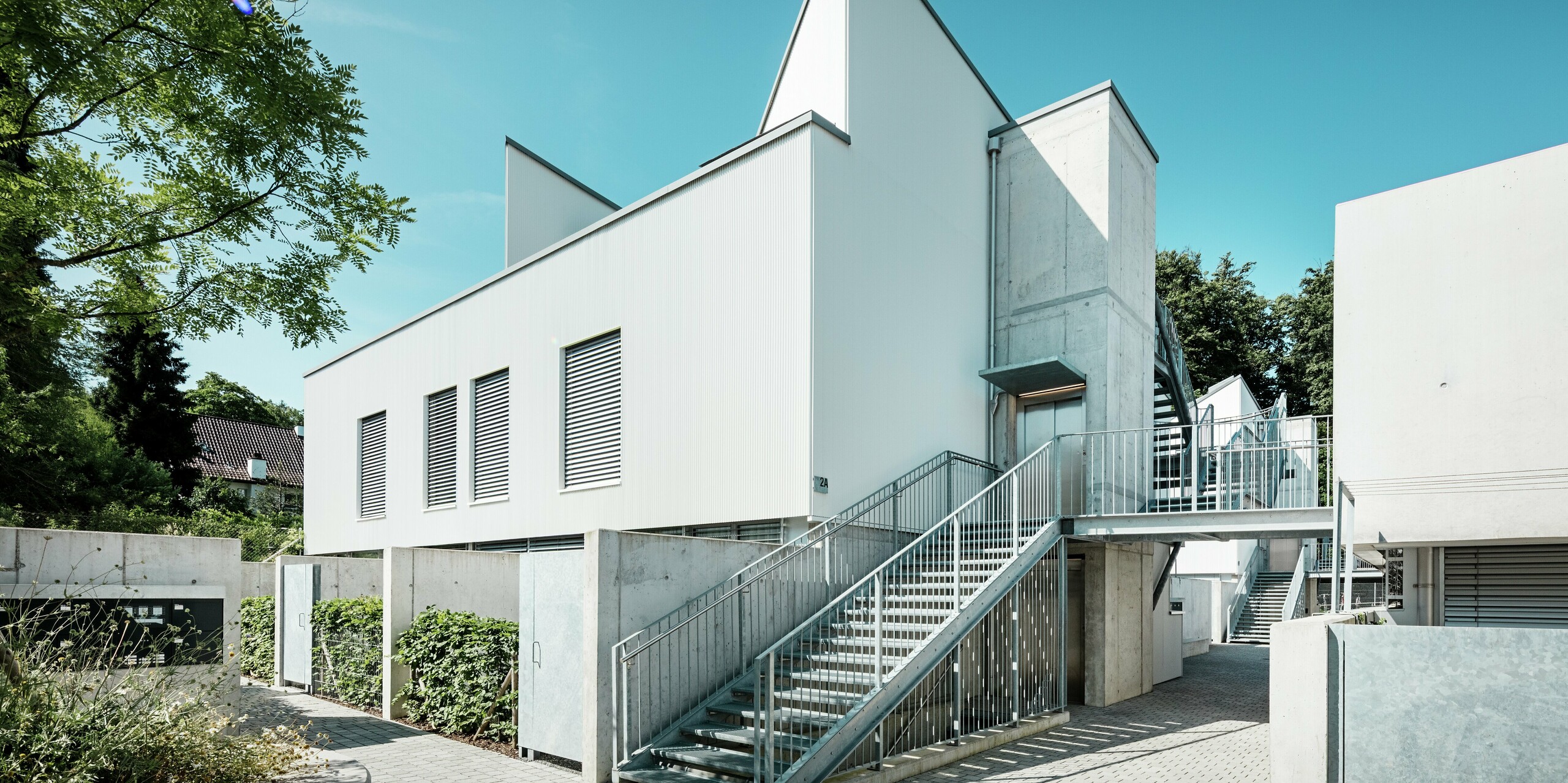 Un ensemble de mini-maisons modernes aux toits à deux pans remarquables, revêtues de profilés triangle PREFA P.10 Blanc Pur. La façade, simple et épurée, s'harmonise avec les éléments en béton apparent et la structure métallique environnante. Durable, la façade en aluminium PREFA assure une protection fiable contre les intempéries et garantit un aspect minimaliste et esthétique, nécessitant peu d'entretien.