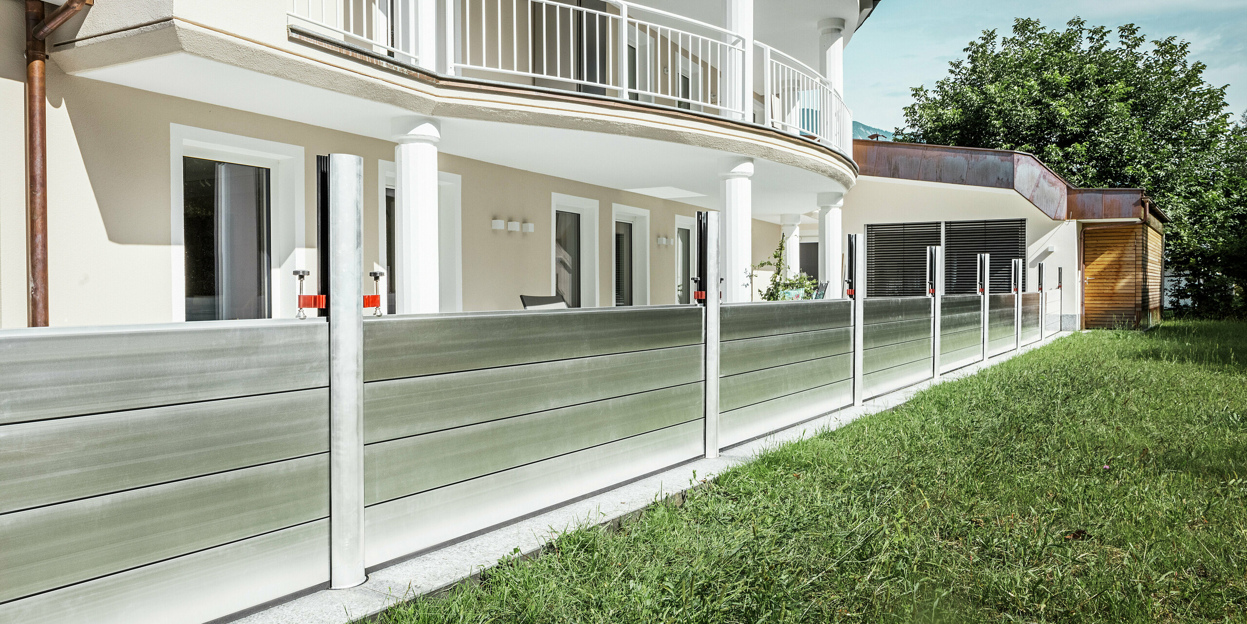 Vue panoramique d'un immeuble résidentiel exclusif au Tyrol, doté d'un système de protection contre les crues PREFA installé le long de la terrasse. Les batardeaux en aluminium s'étendent sur toute la longueur du bâtiment et protègent plusieurs entrées, telles que les portes-fenêtres et les fenêtres, des inondations potentielles provenant de la propriété adjacente. L'architecture moderne de la maison, avec son balcon arrondi et ses colonnes blanches, contraste avec la protection anti-inondation fonctionnelle en aluminium.