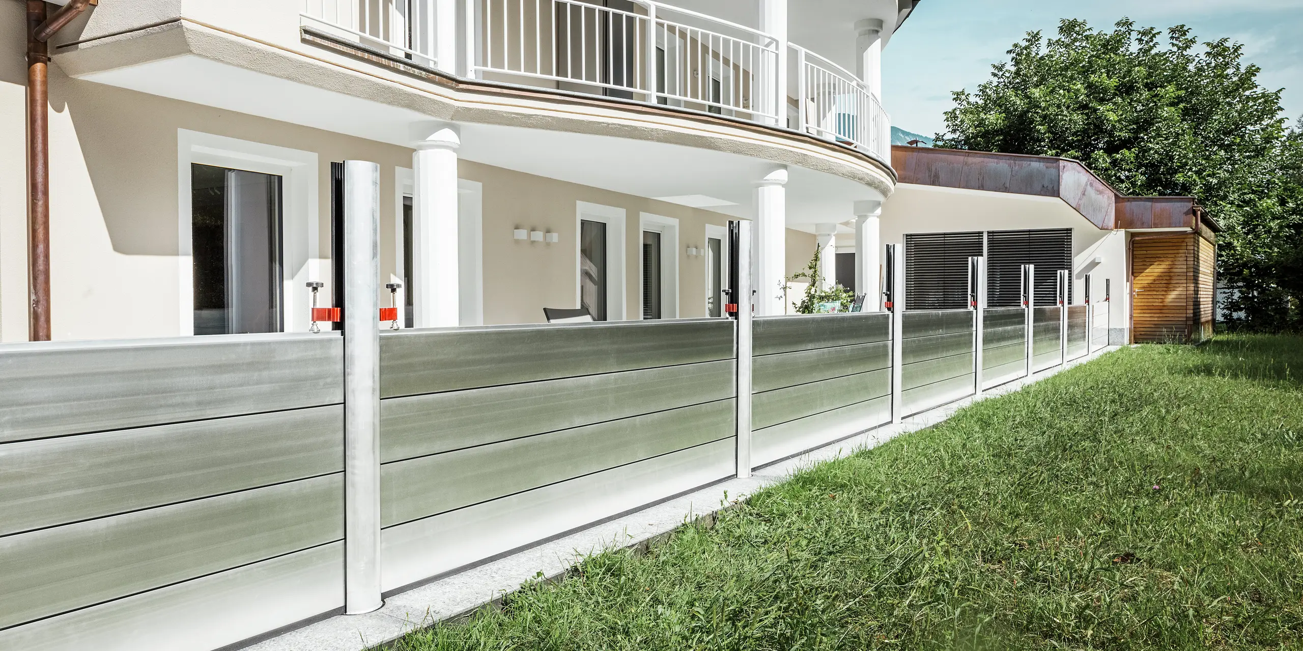 Vue panoramique d'un immeuble résidentiel exclusif au Tyrol, doté d'un système de protection contre les crues PREFA installé le long de la terrasse. Les batardeaux en aluminium s'étendent sur toute la longueur du bâtiment et protègent plusieurs entrées, telles que les portes-fenêtres et les fenêtres, des inondations potentielles provenant de la propriété adjacente. L'architecture moderne de la maison, avec son balcon arrondi et ses colonnes blanches, contraste avec la protection anti-inondation fonctionnelle en aluminium.