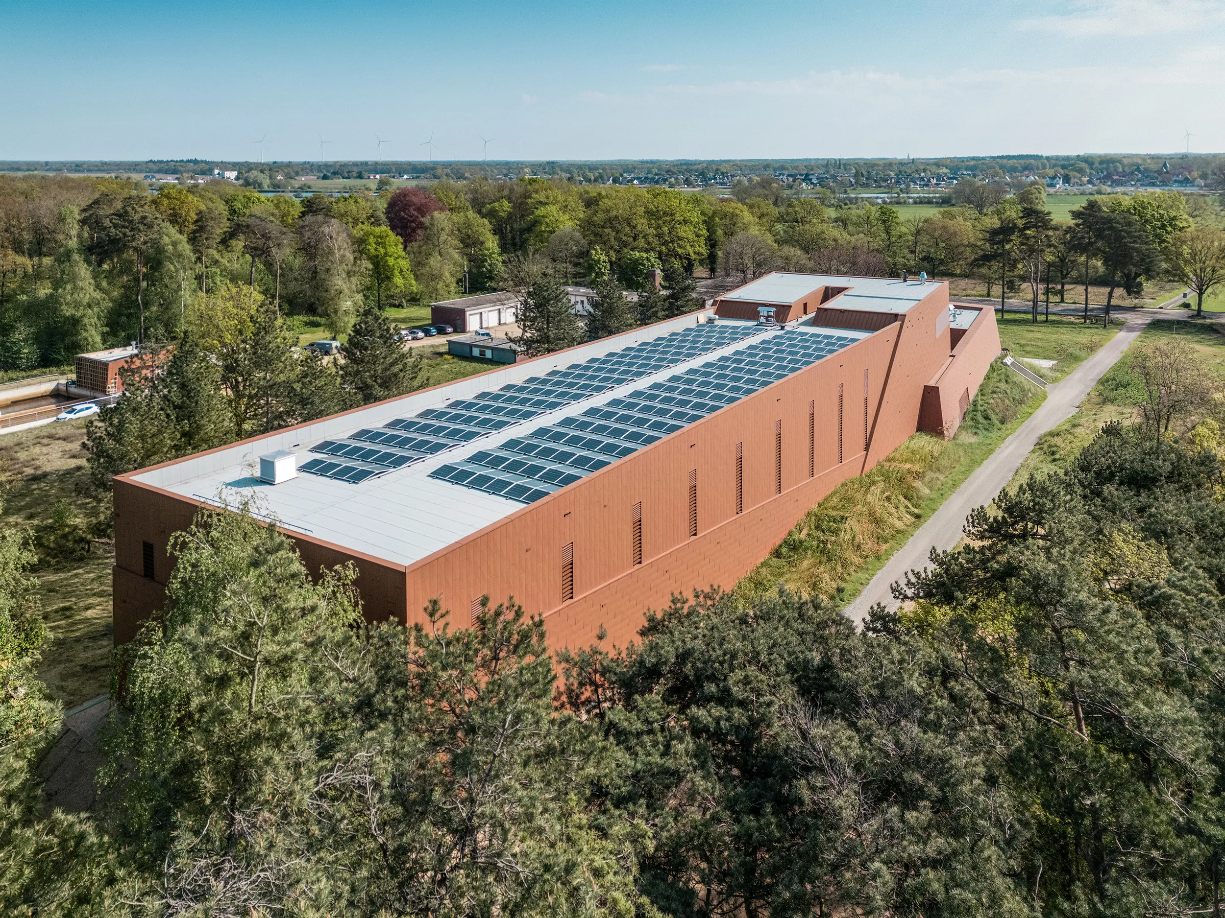 Vue aérienne de la station de pompage d'eau potable WML à Lottum, nichée au cœur d'un vaste paysage arboré et champêtre. La structure allongée du bâtiment impressionne par sa façade en aluminium PREFA Siding.X à la finition chaleureuse et rouillée, et son toit recouvert de modules photovoltaïques, créant ainsi un ensemble architectural durable et remarquable. L'enveloppe du bâtiment, à la structure fine, reflète la lumière avec éclat et souligne la modernité de son design. Légers, durables et recyclables, les panneaux en aluminium PREFA garantissent une excellente résistance aux intempéries, une efficacité énergétique optimale et une architecture pérenne nécessitant peu d'entretien.