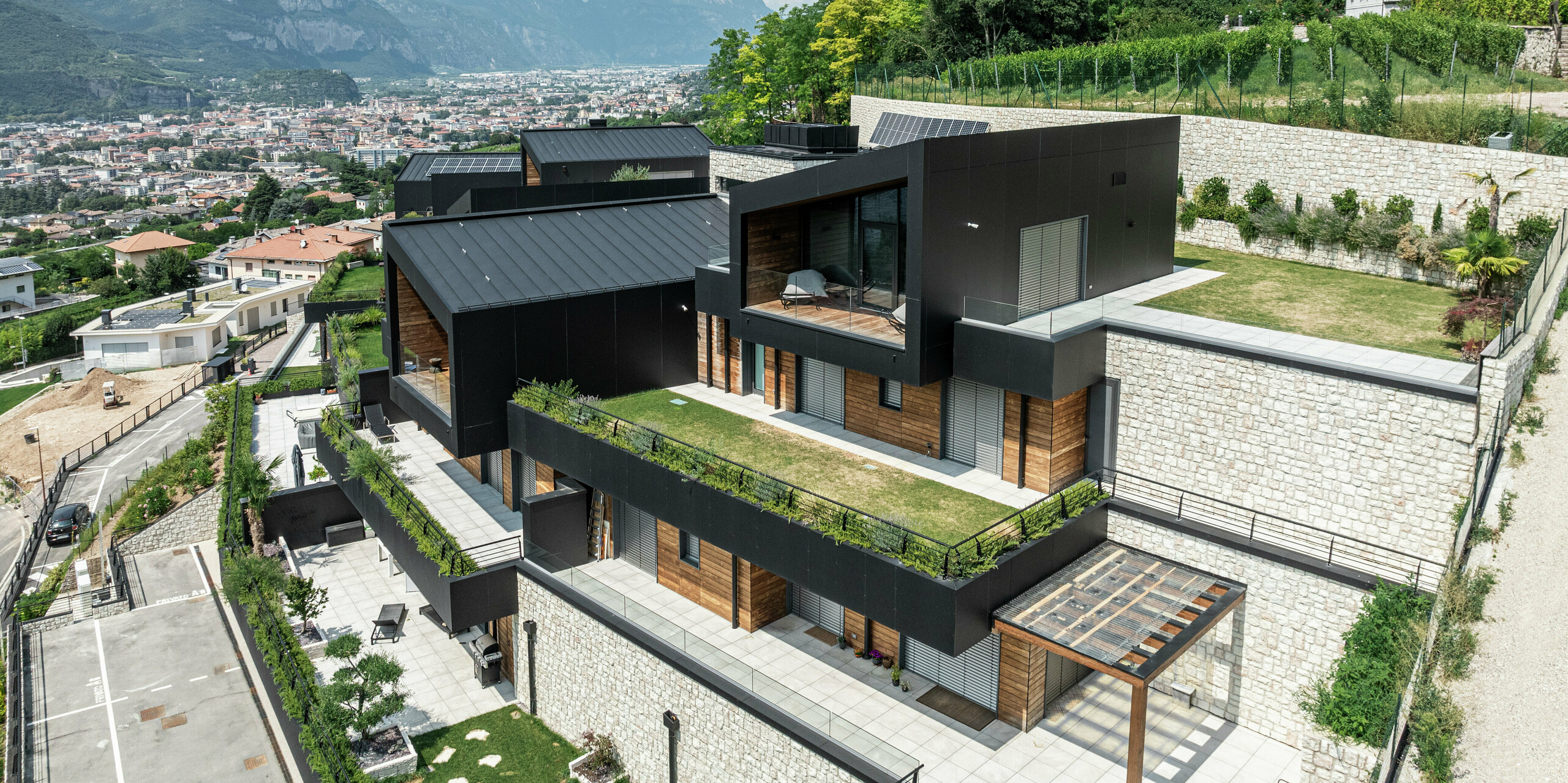 Complexe résidentiel exclusif avec vue panoramique sur Trente : l'architecture moderne s'intègre harmonieusement dans l'emplacement à flanc de colline et combine des matériaux de haute qualité tels que la pierre naturelle, le bois et l'aluminium. La façade en panneaux composites en aluminium PREFA en P.10 Noir assure une apparence élégante et durable, tandis que le toit PREFALZ en P.10 Noir offre une protection optimale contre les éléments. Les espaces verts et les terrasses spacieuses soulignent le concept de vie durable et créent une combinaison parfaite de nature et de design moderne.