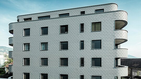 Vue de face de la Bernerhaus à Buchs, avec ses angles arrondis et sa façade continue en bardage PREFA gris souris P.10. La façade en aluminium, à l'aspect d'écailles, crée un motif dynamique et souligne l'horizontalité des balcons incurvés. Les ouvertures, parfaitement intégrées, se fondent harmonieusement dans la surface homogène. Grâce à la légèreté, la résistance aux intempéries et la durabilité de l'aluminium PREFA, l'enveloppe du bâtiment, stable dimensionnellement et nécessitant peu d'entretien, offre un contraste saisissant avec le paysage alpin.
