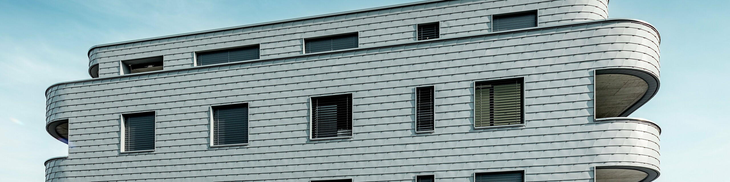 Vue de face de la Bernerhaus à Buchs, avec ses angles arrondis et sa façade continue en bardage PREFA gris souris P.10. La façade en aluminium, à l'aspect d'écailles, crée un motif dynamique et souligne l'horizontalité des balcons incurvés. Les ouvertures, parfaitement intégrées, se fondent harmonieusement dans la surface homogène. Grâce à la légèreté, la résistance aux intempéries et la durabilité de l'aluminium PREFA, l'enveloppe du bâtiment, stable dimensionnellement et nécessitant peu d'entretien, offre un contraste saisissant avec le paysage alpin.
