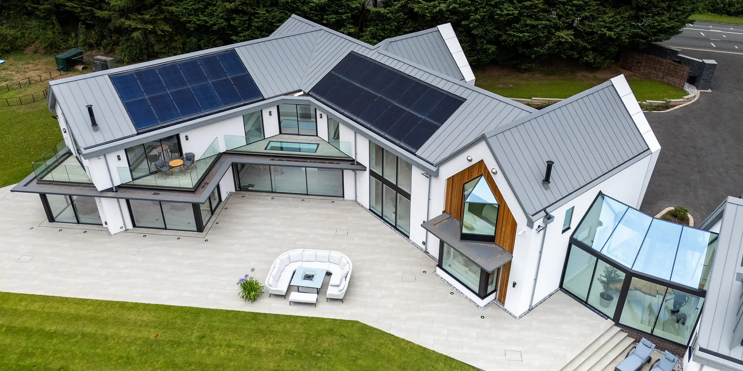 La vue aérienne révèle l'impressionnante villa moderne de Draycott In The Clay, dotée d'une toiture PREFALZ gris quartz. Grâce à sa structure à joints debout épurée et son système photovoltaïque intégré, la toiture allie élégance et haute performance énergétique. La large façade vitrée et le remarquable bardage en bois à l'angle de la maison soulignent la complexité architecturale de la propriété. La vaste terrasse et le jardin paysager créent une harmonie entre le bâtiment et la nature, tandis que la toiture en aluminium PREFALZ garantit une protection et une longévité maximales.