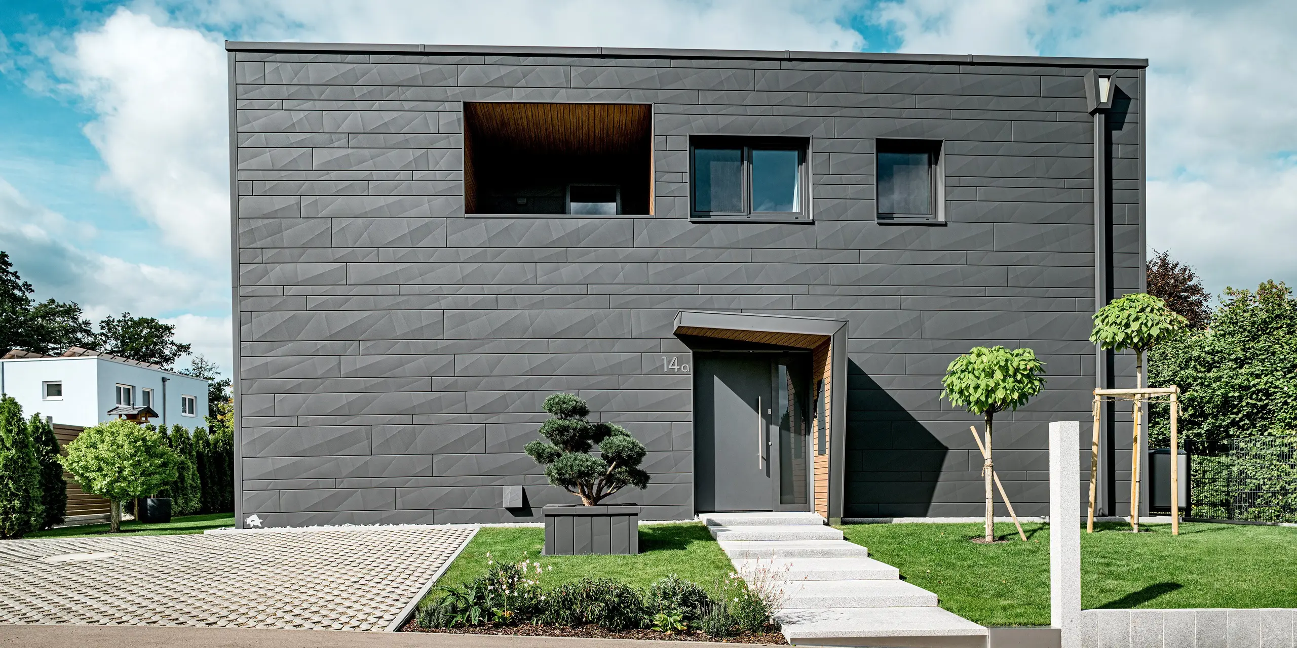Vue de face d'une maison individuelle moderne à Heidenheim, revêtue d'une façade continue en bardage Siding.X PREFA couleur P.10 gris sombre. Les lignes affirmées et les largeurs variables des panneaux créent une façade dynamique. L'entrée habillée de bois et la loggia à l'étage apportent une touche chaleureuse à l'architecture épurée en aluminium. Au premier plan, des topiaires, de jeunes arbres et une cour pavée structurent l'espace. La robuste surface en aluminium PREFA offre une protection durable contre les intempéries et souligne l'aspect moderne et précis du bâtiment.