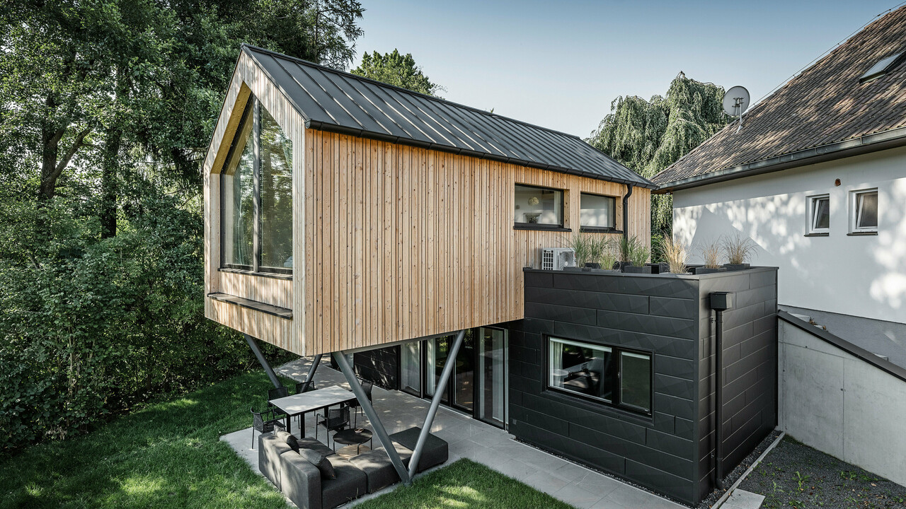 Das 'Woodstock Chalet 5' beeindruckt mit einer eindrucksvollen Kombination aus natürlichen Holzoberflächen und den modernen PREFA Siding.X Fassadenelementen in Anthrazit. Die überhängende Holzstruktur des Obergeschosses ragt markant über die dunklen Fassadenelemente, die das Erdgeschoss verkleiden, und erzeugt so ein Spiel aus Licht und Schatten. Die klaren Linien und die robuste Konstruktion der Siding.X Elemente ergänzen das innovative Design des Gebäudes und bieten einen spannenden visuellen Kontrast zur umgebenden Vegetation und dem traditionellen Gebäude im Hintergrund. Am Dach des Chalets wurde das PREFALZ Dachsystem in Form einer gleichmäßigen Stehfalzdeckung verlegt. Am rechten Rand des Erdgeschosses ist das PREFA Dachentwässerungssystem zu erkennen, das analog zur Aluminiumfassade in Anthrazit gehalten ist. Für die Dachentwässerung wurden der Wasserfangkasten gefolgt vom Ablaufrohr montiert.