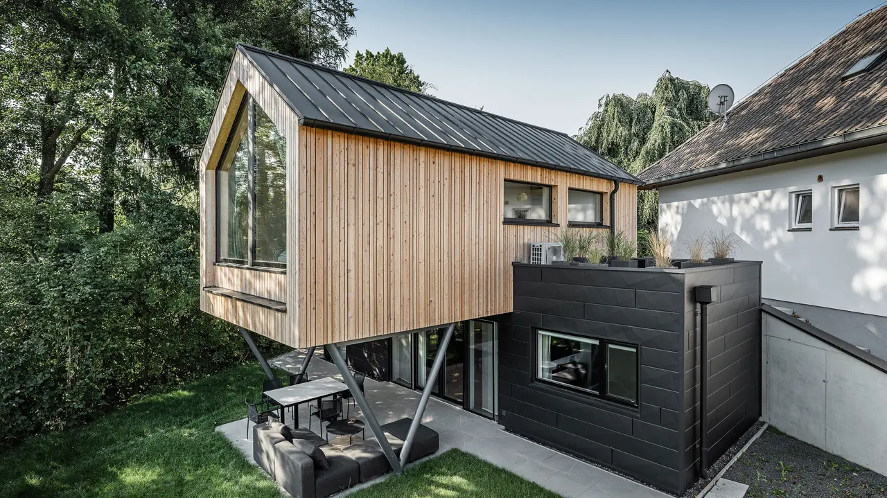 Das 'Woodstock Chalet 5' beeindruckt mit einer eindrucksvollen Kombination aus natürlichen Holzoberflächen und den modernen PREFA Siding.X Fassadenelementen in Anthrazit. Die überhängende Holzstruktur des Obergeschosses ragt markant über die dunklen Fassadenelemente, die das Erdgeschoss verkleiden, und erzeugt so ein Spiel aus Licht und Schatten. Die klaren Linien und die robuste Konstruktion der Siding.X Elemente ergänzen das innovative Design des Gebäudes und bieten einen spannenden visuellen Kontrast zur umgebenden Vegetation und dem traditionellen Gebäude im Hintergrund. Am Dach des Chalets wurde das PREFALZ Dachsystem in Form einer gleichmäßigen Stehfalzdeckung verlegt. Am rechten Rand des Erdgeschosses ist das PREFA Dachentwässerungssystem zu erkennen, das analog zur Aluminiumfassade in Anthrazit gehalten ist. Für die Dachentwässerung wurden der Wasserfangkasten gefolgt vom Ablaufrohr montiert.