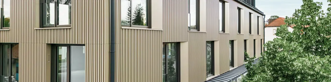 La photo montre un bâtiment moderne à Feldkirchen, conçu par l'architecte Stefan Heigl. La façade est en Prefalz de couleur bronze et le toit à pignon est recouvert de Prefalz de couleur gris noir. Les grandes fenêtres apportent beaucoup de lumière à l'intérieur, tandis que le rez-de-chaussée vitré offre un coin repas accueillant. Le choix des matériaux de haute qualité et le design élégant confèrent au bâtiment un aspect contemporain et élégant.