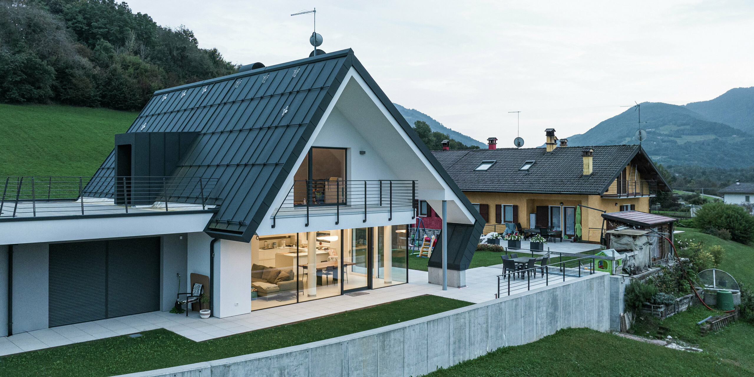 Maison individuelle moderne avec vue sur jardin et terrasse. Le toit en aluminium avec des surplombs marquants protège la façade et crée une transition harmonieuse vers la terrasse spacieuse. De grandes fenêtres assurent des pièces lumineuses et le balcon enveloppant souligne le caractère ouvert du salon. La combinaison de l'aluminium PREFA durable, de l'architecture moderne et de la finition de haute qualité rend cette maison particulièrement durable et élégante.