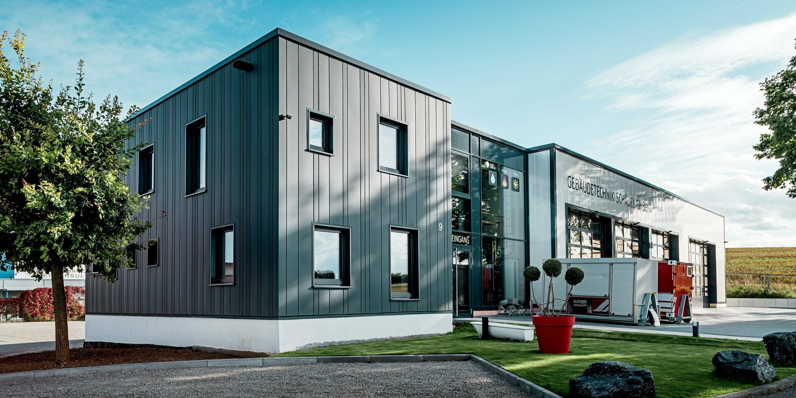 Die Szene zeigt den klar geformten Baukörper der Gebäudetechnik Schädel GmbH mit seiner prägnanten PREFA Blechfassade, deren vertikale Linien in Anthrazit sofort ins Auge fallen. Die Mischung aus verschiedenen Siding-Breiten verleiht dem Gebäude einen rhythmischen Charakter. Das Zusammenspiel aus Aluminium, Glasfront und offenem Vorplatz schafft eine moderne, kraftvolle Wirkung. Die langlebige PREFA Oberfläche bewährt sich gegen Wetter und Abnutzung.