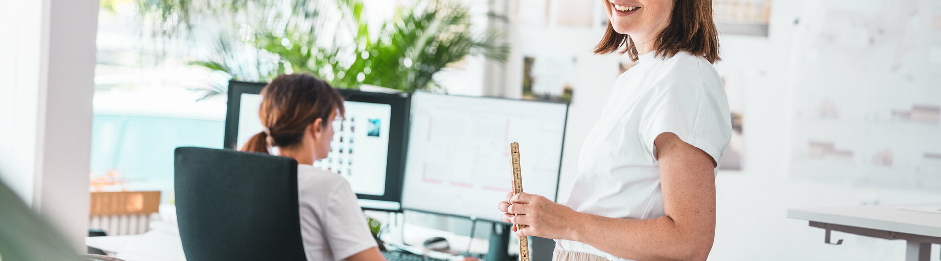Zwei Architektinnen im modernen Büro: Eine arbeitet am Computer mit digitalen Plänen, während die andere mit Maßstab und Entwürfen bereitsteht. Das Bild steht für effizienten Zugriff auf Planungsunterlagen und technische Ressourcen im PREFA Downloadcenter für Architekten und Planer.