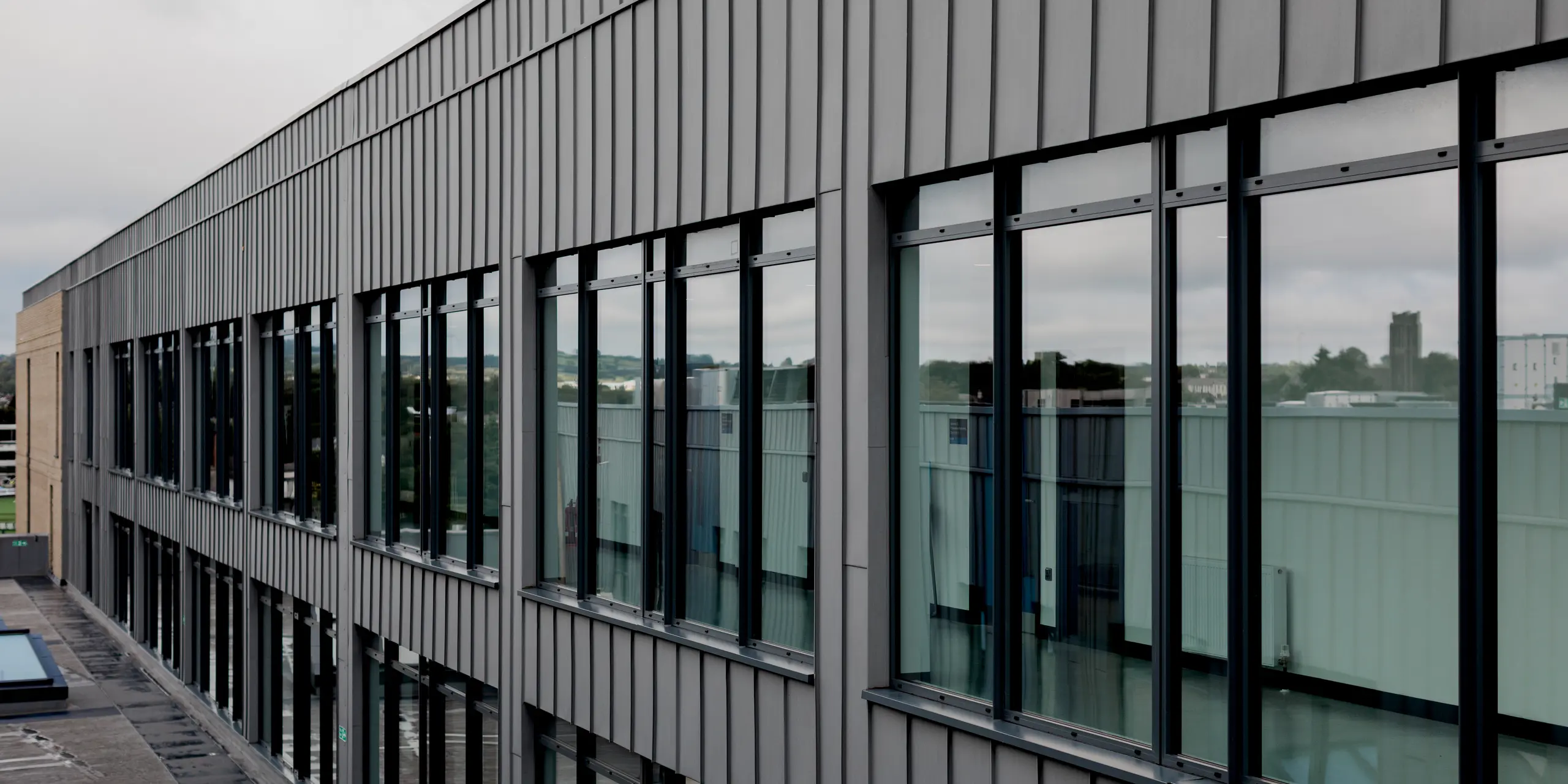 La longue et élégante façade du Northern Regional College de Ballymena présente un revêtement continu en aluminium PREFALZ gris quartz, à joints debout parfaitement verticaux. De larges fenêtres régulièrement espacées reflètent le ciel et le paysage environnant, conférant au bâtiment transparence et profondeur. Le revêtement métallique forme un cadre sobre et épuré pour les surfaces vitrées et souligne la linéarité de l'architecture. L'aluminium PREFA impressionne ici par sa précision, sa durabilité, sa résistance aux intempéries et son esthétique moderne et haut de gamme.