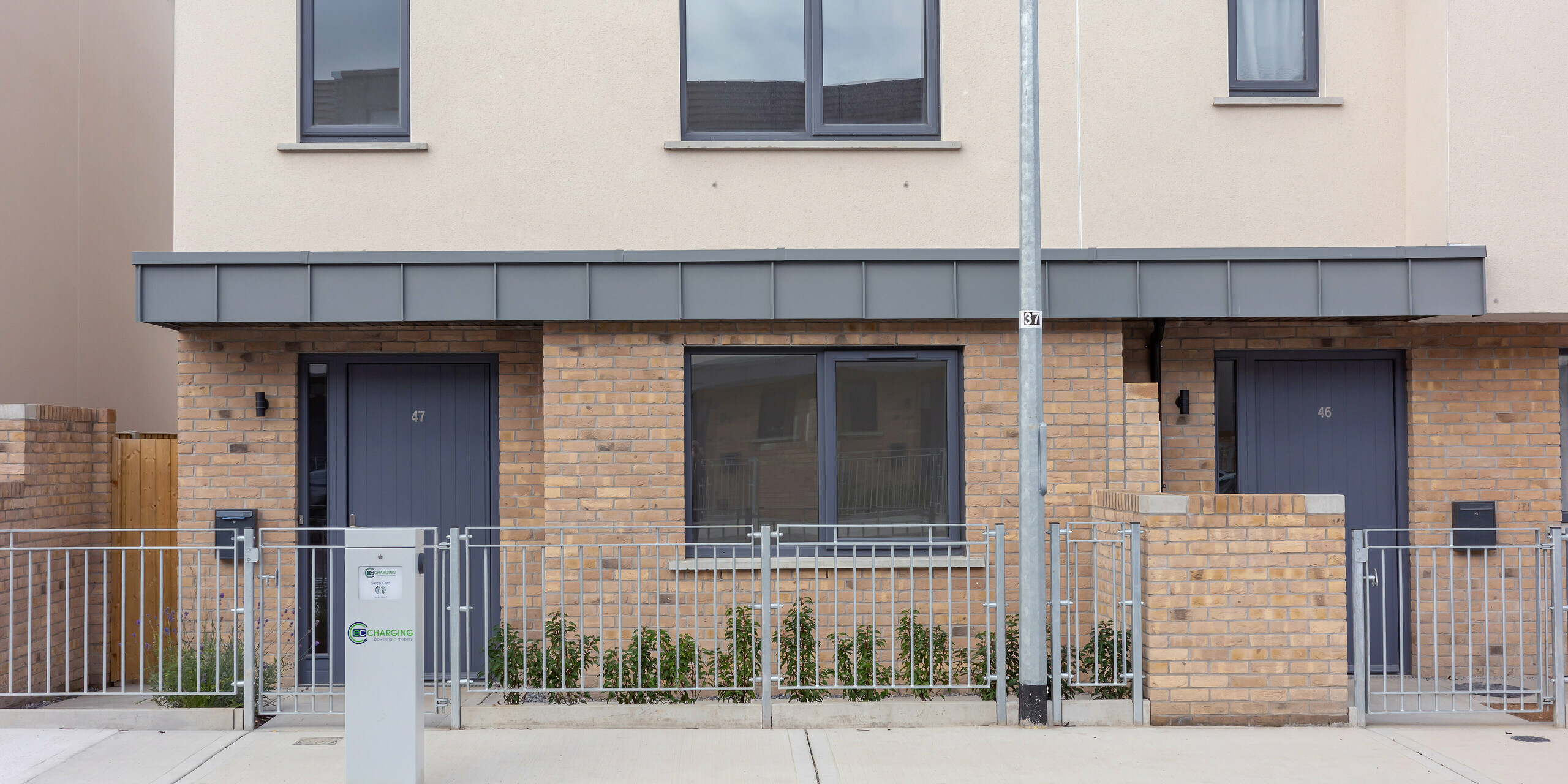 L'image présente l'entrée d'un ensemble de maisons mitoyennes modernes à Churchfields, caractérisé par des lignes épurées et des couleurs harmonieuses. L'élégant auvent au-dessus des portes d'entrée, revêtu de PREFALZ P.10 gris souris, est particulièrement remarquable. La robuste surface en aluminium apporte une touche de modernité à la chaleureuse maçonnerie de briques et ne nécessite pratiquement aucun entretien – un atout majeur pour les grands projets résidentiels. L'homogénéité de la façade et le jardin soigné à l'avant confèrent à l'ensemble une atmosphère sereine.
