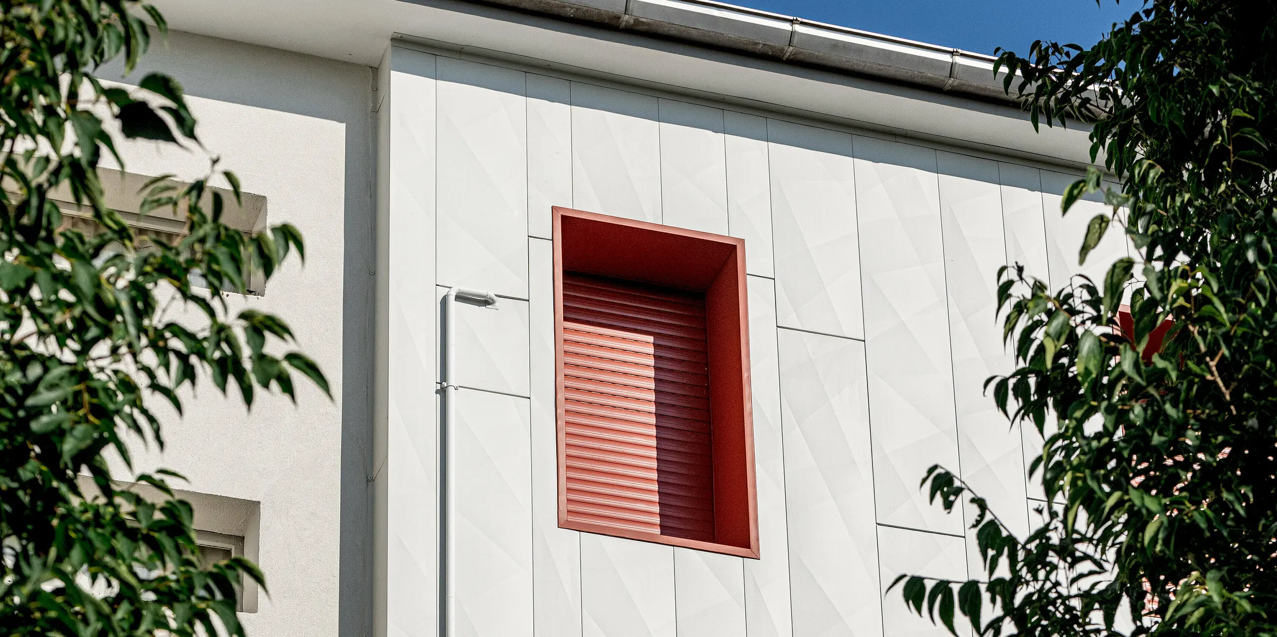 Le complexe résidentiel moderne Alba B, après rénovation avec façade en aluminium PREFA Siding.X, présente un jeu d'ombres et de lumières captivant, grâce à ses panneaux verticaux. Les encadrements de fenêtres rouges contrastent fortement avec la façade, attirant le regard. Entouré de cimes verdoyantes, le bâtiment dégage une impression de dynamisme et de sophistication. La façade en aluminium PREFA séduit par sa durabilité, sa résistance et son faible entretien, tout en offrant une esthétique contemporaine et architecturale distinctive.