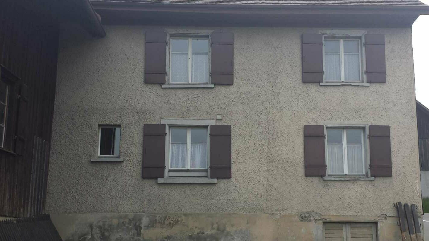 Maison avant la rénovation de façade à l’aide de losanges de façade PREFA, fenêtres dotées de volets rouges