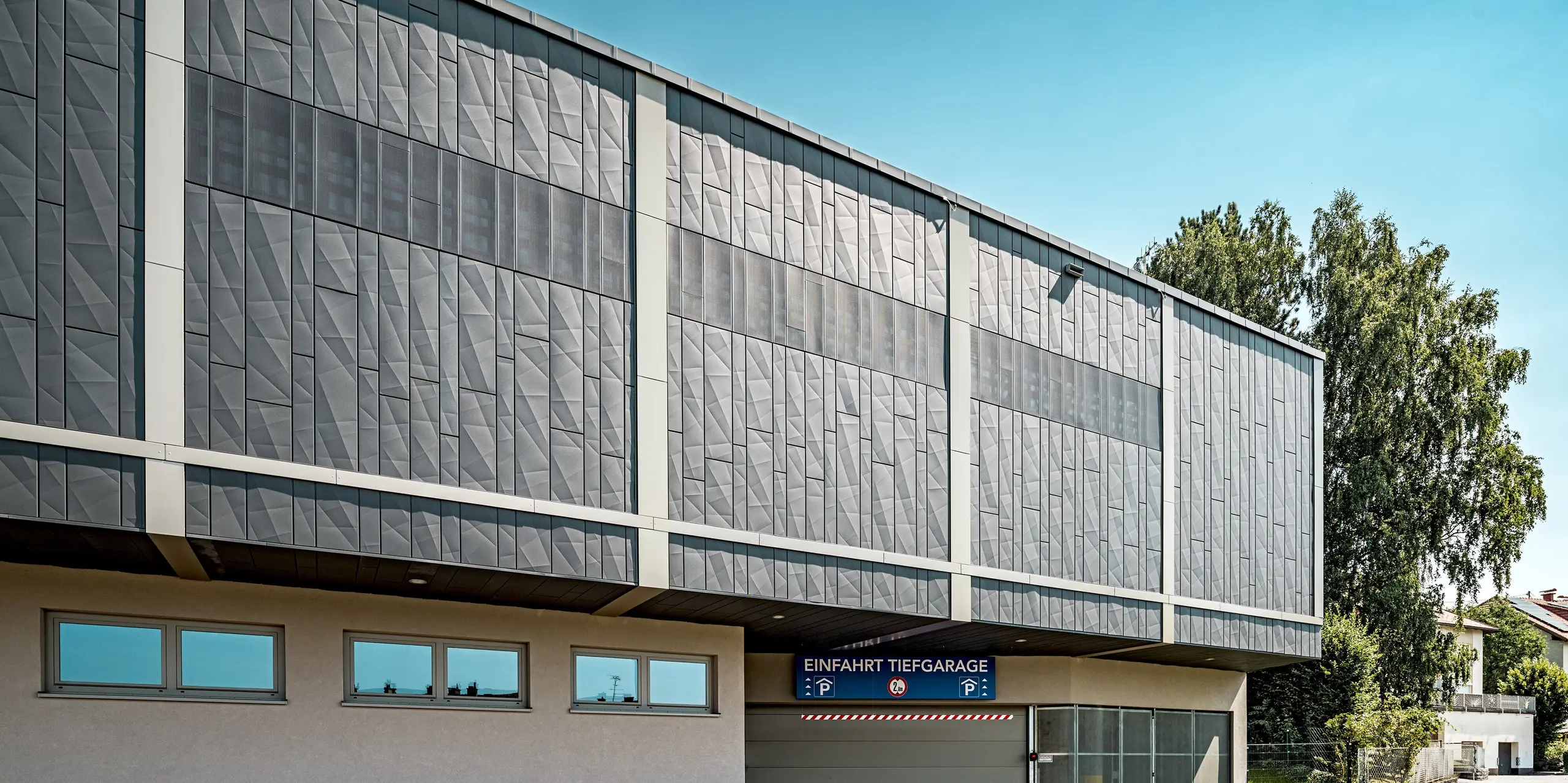 L'image montre l'entrée du parking souterrain du centre commercial de Rohrbach. Le bâtiment est revêtu de Siding.X PREFA anthracite. Les éléments de façade à la structure géométrique sont particulièrement remarquables ; ils confèrent au bâtiment une allure moderne et s'harmonisent parfaitement avec l'entrée du parking. La pureté des lignes architecturales est accentuée par l'emplacement précis des fenêtres et le bardage perforé.