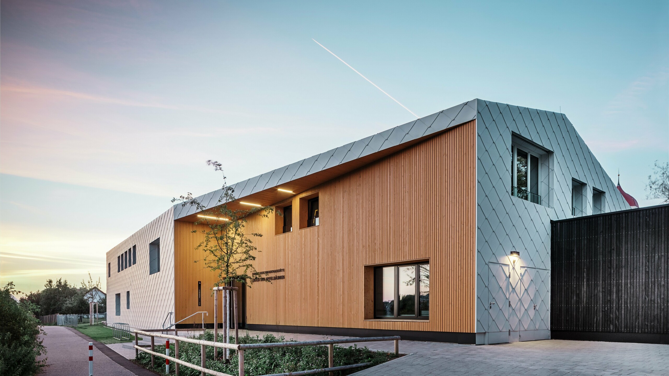 Vue d'une crèche moderne à Ulm au crépuscule. La toiture a été réalisée sans avant-toit. La majeure partie de la façade a été habillée de grands losanges en aluminium blanc de PREFA, tandis que la zone d'entrée a été habillée de grands panneaux de bois verticaux.