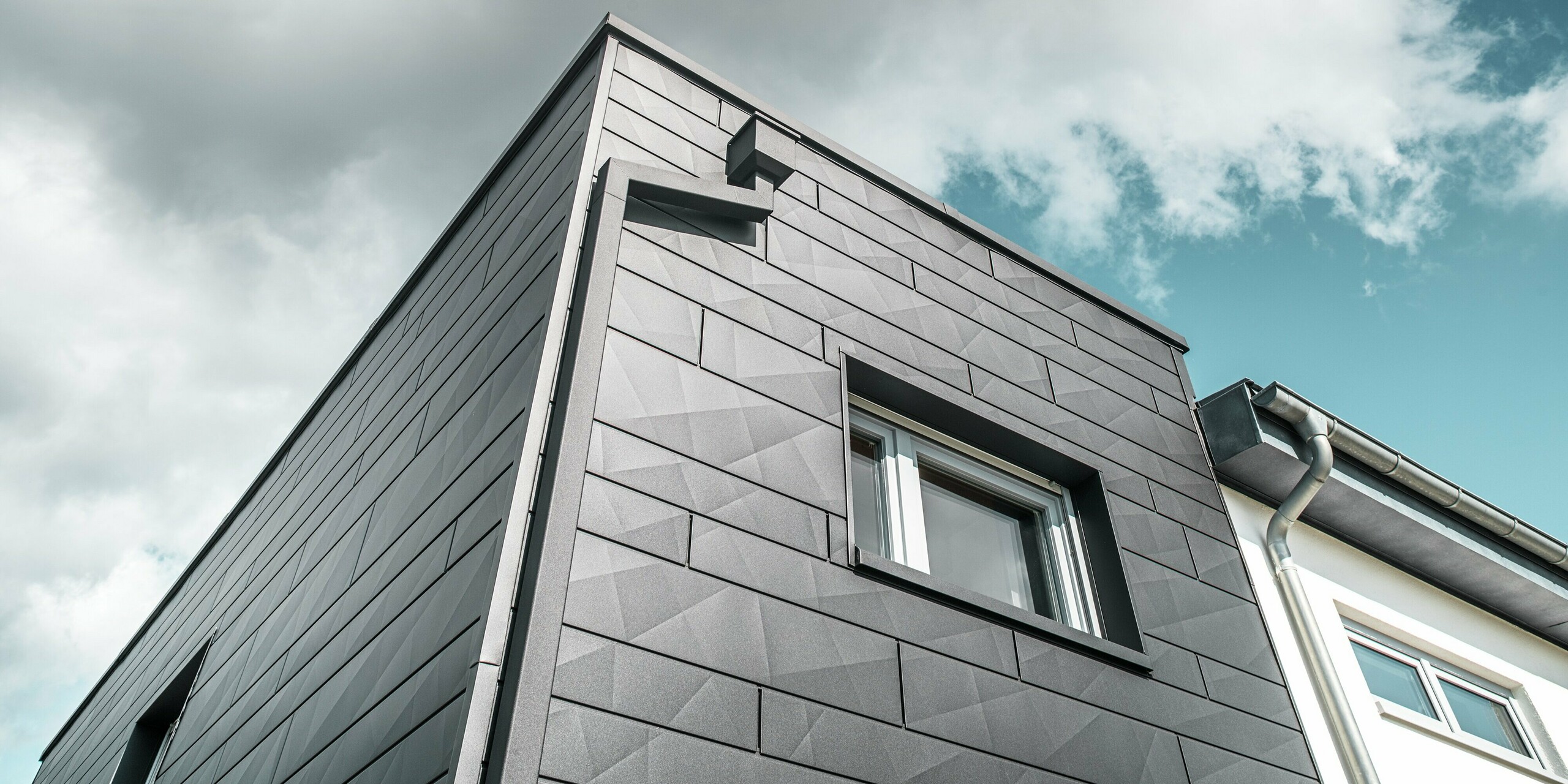 La façade remarquable de la maison individuelle rénovée de Teuschnitz est ornée de panneaux en aluminium Siding.X PREFA couleur P.10 noir, créant une structure moderne et géométrique. Les éléments de façade suivent des lignes épurées et soulignent le caractère intemporel du bâtiment. Une fenêtre est encadrée de panneaux composites en aluminium noir. Le système intégré de gouttière PREFA, avec son tuyau carré et sa boîte à eau, s'intègre harmonieusement au bâtiment et offre une solution élégante et fonctionnelle.