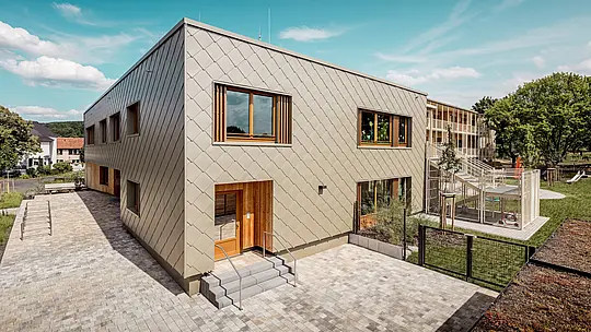 L'image présente la crèche municipale d'Alzenau, vue en plongée oblique, mettant en valeur son architecture moderne et sa remarquable façade en aluminium PREFA couleur bronze. Cette façade est composée de losanges de façade de 44 x 44 cm, créant un motif harmonieux évoquant des écailles et conférant au bâtiment un dynamisme élégant. De grandes fenêtres en bois à persiennes verticales apportent une touche chaleureuse et laissent entrer abondamment la lumière naturelle. À droite de l'image, le jardin s'ouvre sur une aire de jeux clôturée, des espaces verts et une terrasse couverte. L'alliance de l'aluminium, matériau durable et sans entretien, et de matériaux écologiques fait de ce bâtiment un exemple parfait d'architecture fonctionnelle et écologique dans le domaine de la construction éducative.