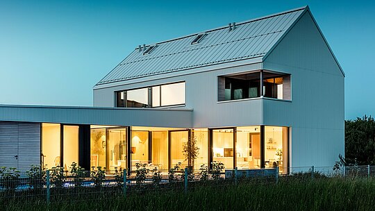 L'image présente une maison individuelle moderne à Freistadt, offrant une perspective qui met en valeur l'harmonie entre architecture et nature. La façade PREFALZ d'un blanc immaculé attire immédiatement le regard, tandis que les larges baies vitrées créent un intérieur ouvert et accueillant. La toiture à forte pente, également revêtue d'aluminium PREFALZ, apporte une touche de dynamisme et protège le bâtiment des intempéries. Le design élégant et minimaliste est rehaussé par les éléments en bois chaleureux du balcon et de la terrasse, créant un contraste agréable. Grâce à la durabilité et à la facilité d'entretien des produits en aluminium PREFALZ, le bâtiment est non seulement esthétique, mais aussi fonctionnel et durable.