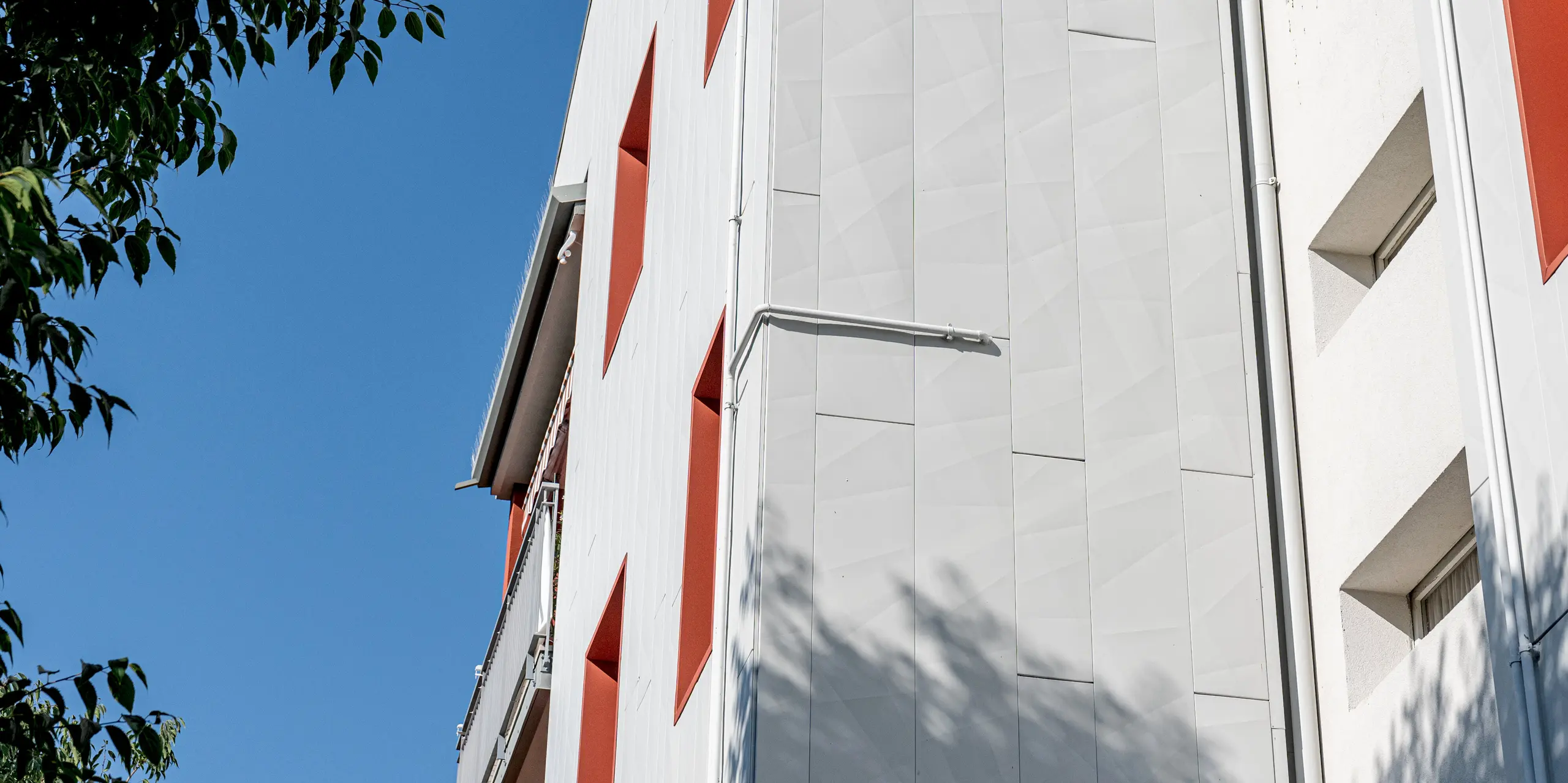 La photographie présente le complexe résidentiel Alba B, modernisé, sous un angle dynamique et en contre-plongée, encadré par des branches d'arbres verdoyantes et un ciel d'un bleu limpide. La façade lumineuse et spacieuse, composée de panneaux en aluminium PREFA Siding.X, dégage une impression à la fois de sérénité et de dynamisme : la finesse des bords et la verticalité de la pose créent de subtils jeux d'ombre et de lumière. Les ouvertures de fenêtres profondément encastrées, avec leurs encadrements rouges audacieux, sont particulièrement saisissantes et créent de forts contrastes. Ici, les produits en aluminium PREFA, durables, résistants aux intempéries et nécessitant peu d'entretien, allient une architecture distinctive à une protection de façade pérenne.