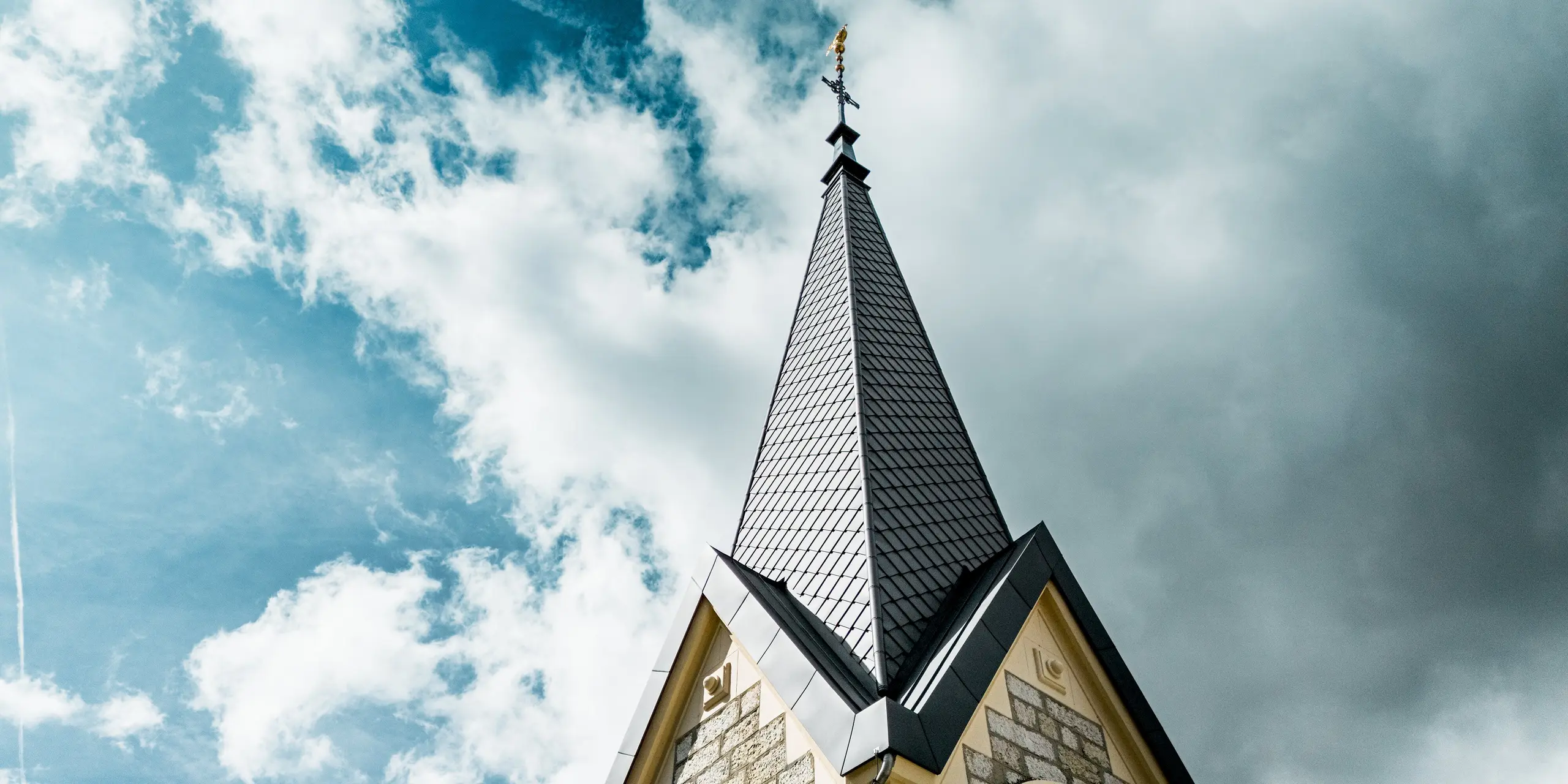Gros plan spectaculaire sur le clocher rénové de l'église du Sentier, en Suisse. Le losange PREFA 29 x 29 cm, coloris Gris sombre P.10, confère à la flèche élancée une structure élégante et précise, et la fait ressortir avec élégance sur le ciel nuageux. Cette toiture en aluminium durable impressionne par sa grande résistance aux intempéries, sa légèreté et son adaptation optimale aux toitures à forte pente.