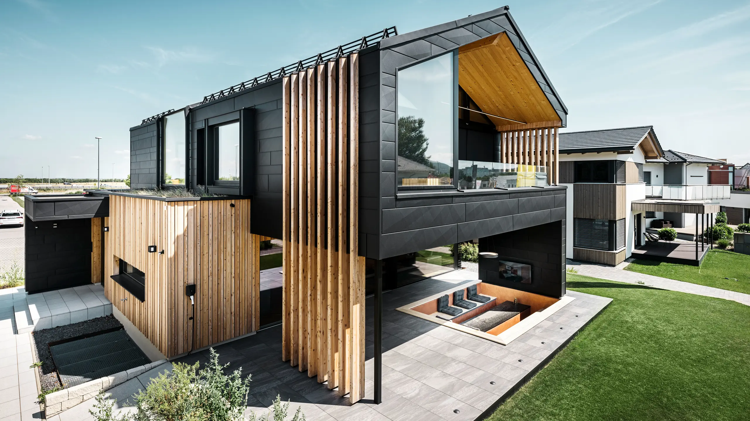 Vue latérale de la maison modèle Autark dans le Blue Lagoon à Vösendorf, en Autriche. L'étage supérieur impressionne par ses grandes surfaces vitrées et sa façade moderne en aluminium composée de panneaux PREFA Siding.X en P.10 noir, associés à des éléments verticaux en bois. La terrasse spacieuse au rez-de-chaussée avec sièges encastrés s'intègre parfaitement dans l'architecture. Le design élégant et durable de la maison combine de manière impressionnante l'aluminium et le bois.
