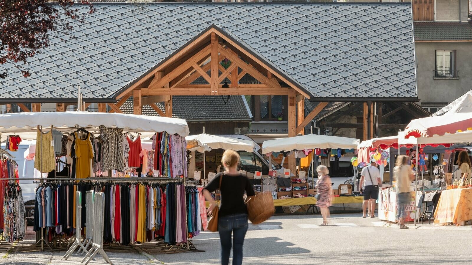 Vue éloignée de la halle couverte d'Allevard, revêtue de losange de toiture 44x44 teinte P.10 brun noisette. On aperçoit le marché et les clients en premier plan.