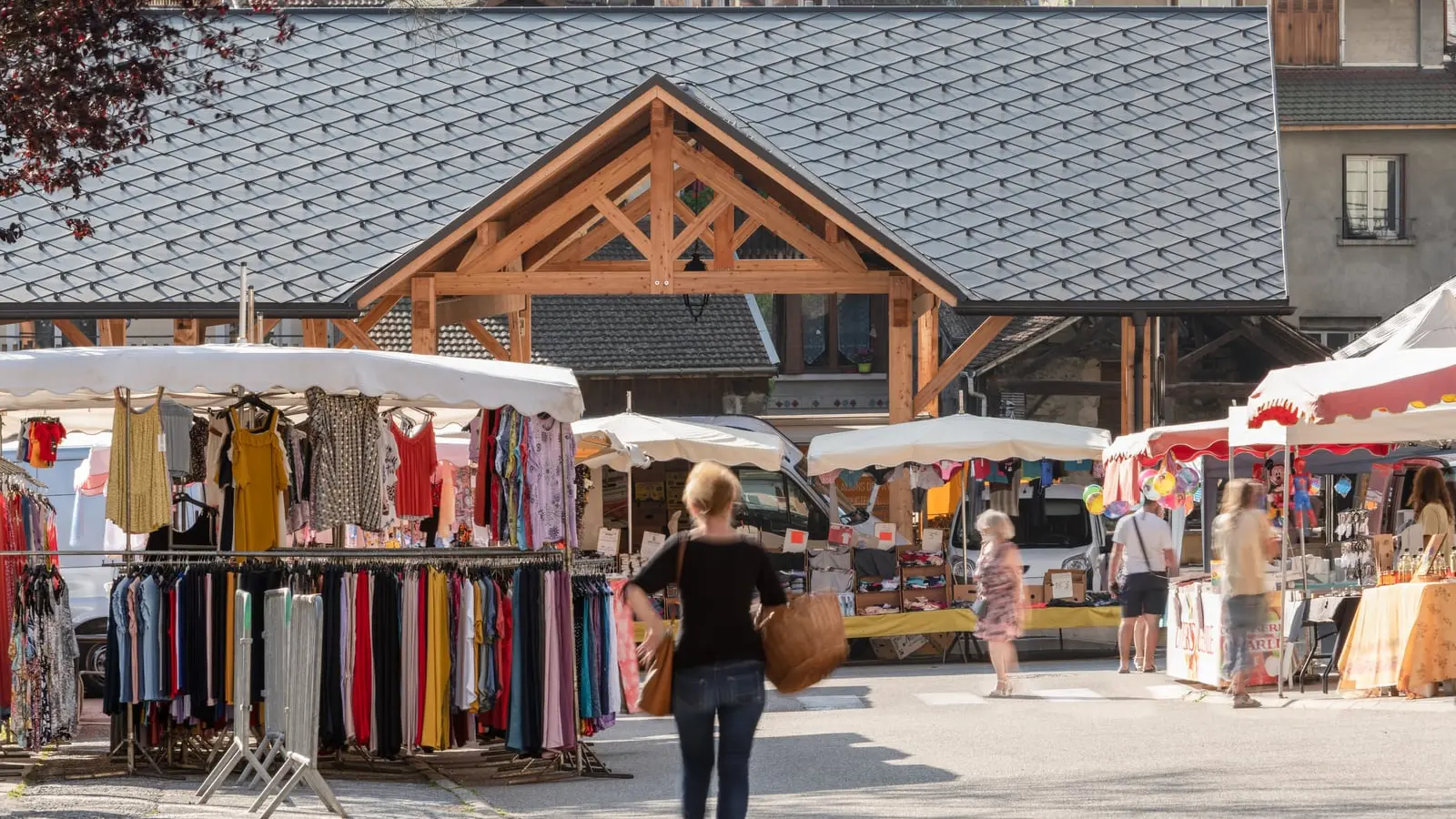 Vue éloignée de la halle couverte d'Allevard, revêtue de losange de toiture 44x44 teinte P.10 brun noisette. On aperçoit le marché et les clients en premier plan.