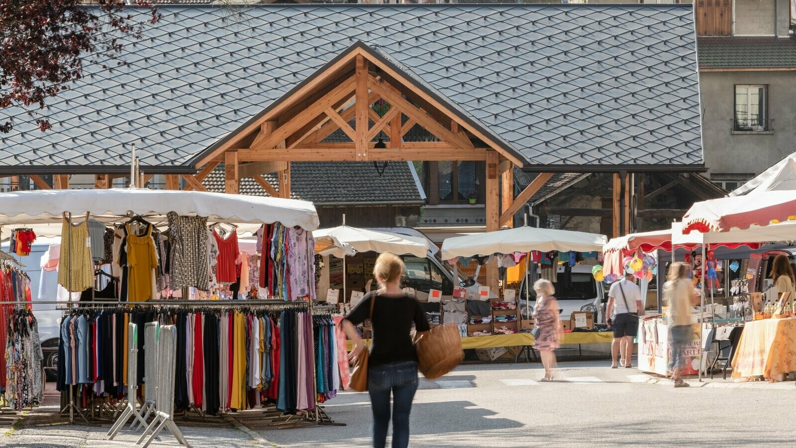 Vue éloignée de la halle couverte d'Allevard, revêtue de losange de toiture 44x44 teinte P.10 brun noisette. On aperçoit le marché et les clients en premier plan.