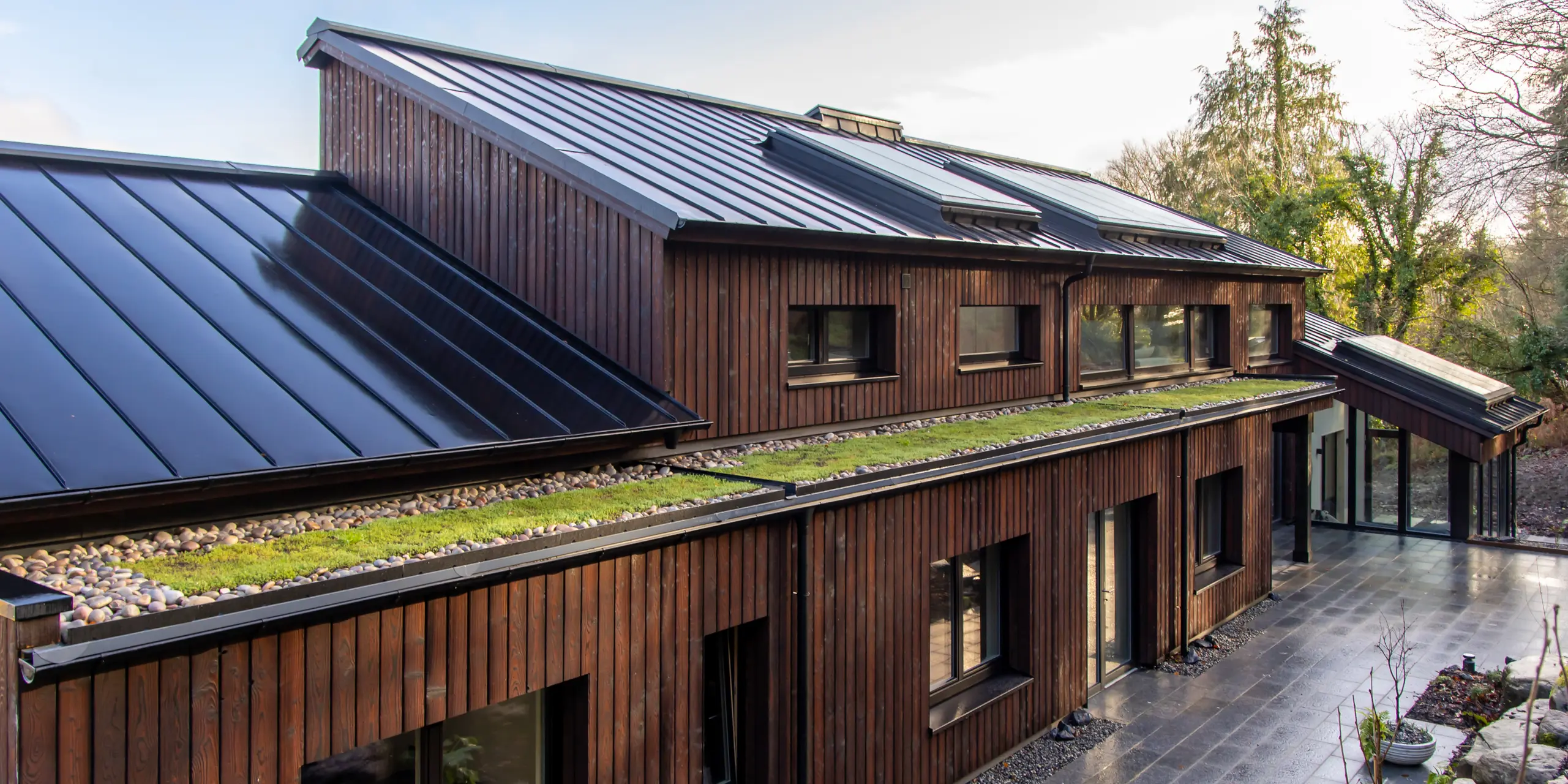 Vue en perspective de la terrasse de la Maison Holywell à Sligo : les surfaces de toiture aux courbes dynamiques, réalisées en aluminium PREFALZ noir P.10, attirent le regard sur toute la longueur du bâtiment. Les joints debout précis structurent la surface et reflètent la lumière, soulignant ainsi la pureté des lignes architecturales. L’association de la toiture sombre, de la façade en bois et des éléments végétaux crée une impression d’ensemble saisissante. L’aluminium PREFA impressionne par sa durabilité, sa résistance à la corrosion et sa protection fiable par tous les temps.