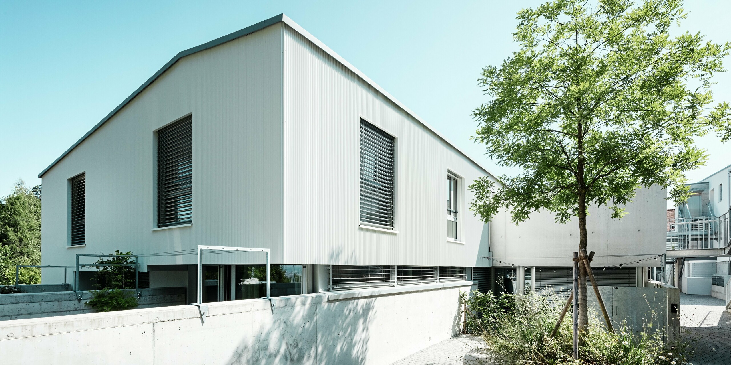 De charmantes mini-maisons modernes à Zollikerberg, en Suisse, arborent une toiture PREFALZ gris quartz et une façade en profils triangle PREFA blanc pur P.10. L'architecture épurée est mise en valeur par des bandeaux de fenêtres horizontaux, des stores vénitiens extérieurs et des éléments en béton apparent. L'aluminium de haute qualité séduit par sa résistance aux intempéries, son faible entretien et son élégance intemporelle. Les arbres qui agrémentent le complexe résidentiel créent une atmosphère de vie paisible.