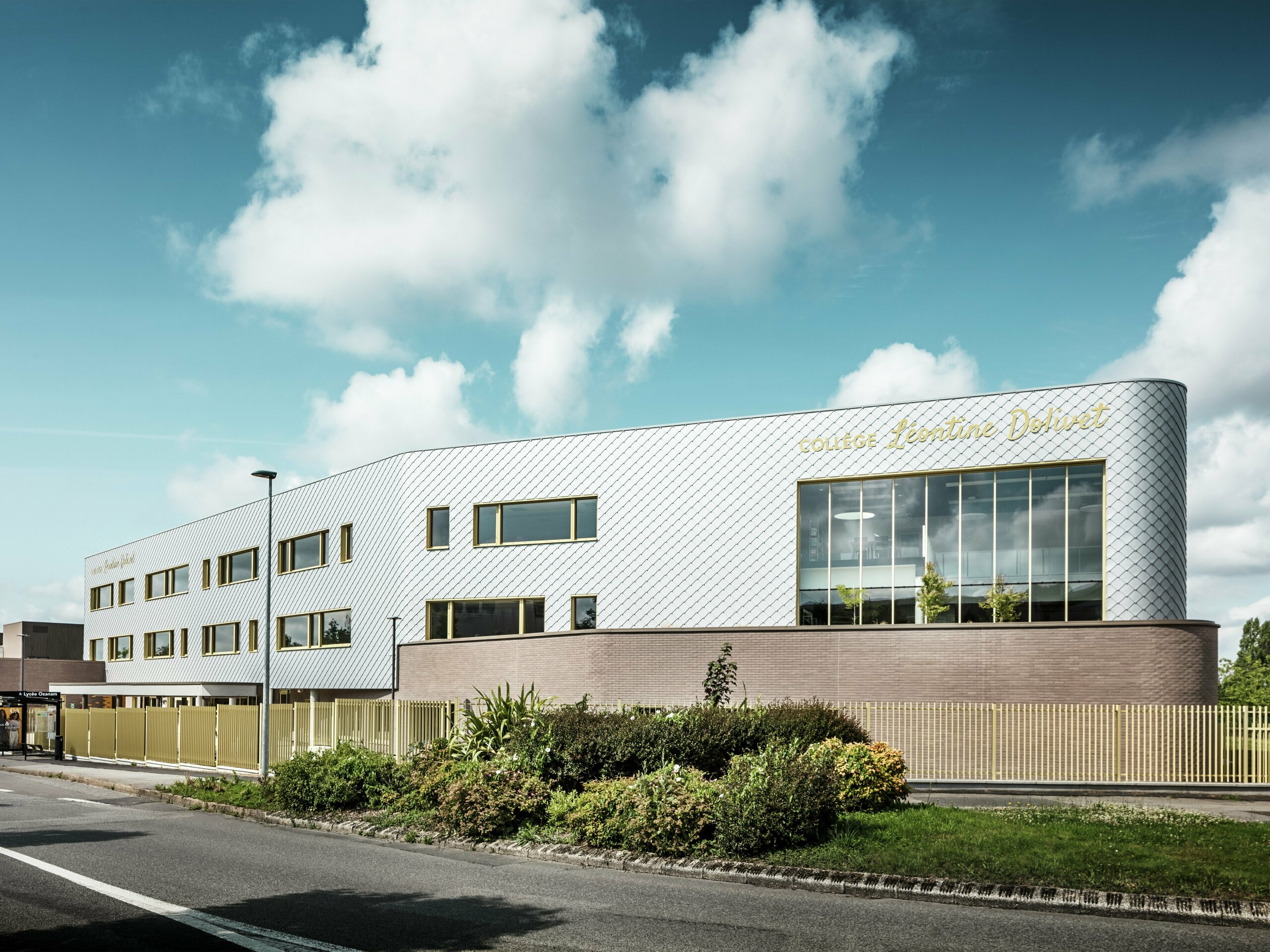 Vue du Collège Léontine Dolivet et de sa façade en aluminium aux reflets argentés, composée de losanges PREFA 29 x 29 couleur argent métallisé. Le motif en losanges diagonaux confère à la façade un aspect dynamique, tandis que de larges surfaces vitrées et des volets dorés créent des contrastes saisissants. Au premier plan, des espaces verts soignés et une clôture moderne encadrent le bâtiment. Solution de façade durable, le système PREFA allie élégance, résistance aux intempéries et durabilité – un véritable joyau architectural à Cesson-Sévigné, près de Rennes.