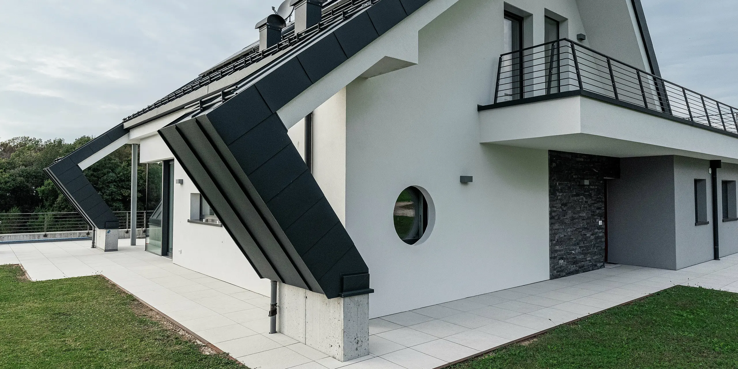 Gros plan de l'extension du toit à pignon d'une maison individuelle moderne à Borgo Valbelluna avec une couverture à joint debout PREFALZ en P.10 anthracite. Le surplomb marquant du toit en aluminium souligne l'architecture innovante et protège la façade. La couverture de toit durable et résistante aux intempéries s'harmonise parfaitement avec la façade en plâtre blanc et les accents sombres. Un balcon enveloppant et des détails de fenêtres rondes confèrent au bâtiment une touche spéciale. L'aluminium PREFA de haute qualité garantit stabilité, élégance et absence d'entretien.