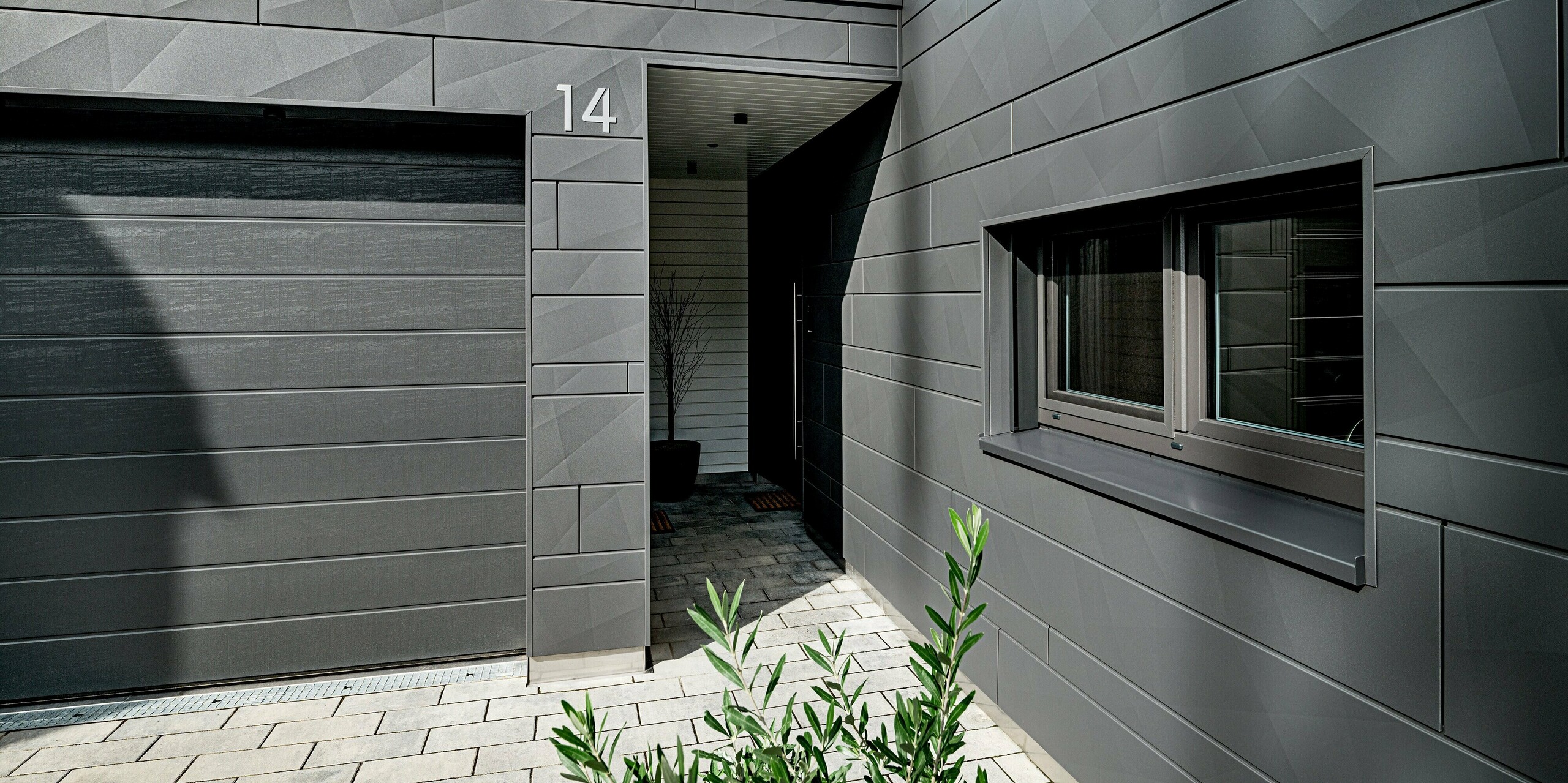 Eingangs- und Garagenbereich eines modernen Einfamilienhauses in Heidenheim mit vollständig verkleideter PREFA Siding.X Aluminiumfassade in P.10 Dunkelgrau. Die markanten Kantstrukturen der Paneele erzeugen ein lebendiges, geometrisches Muster. Die Hausnummer „14“ steht klar sichtbar auf dem Fassadenelement über dem überdachten Zugang. Rechts prägt ein großes, präzise eingefasstes Fenster die Szene, während die gepflasterte Zufahrt und dezente Bepflanzung den modernen Gesamteindruck abrunden. Die langlebige PREFA Oberfläche bietet hohen Schutz und zeitlose Stabilität.