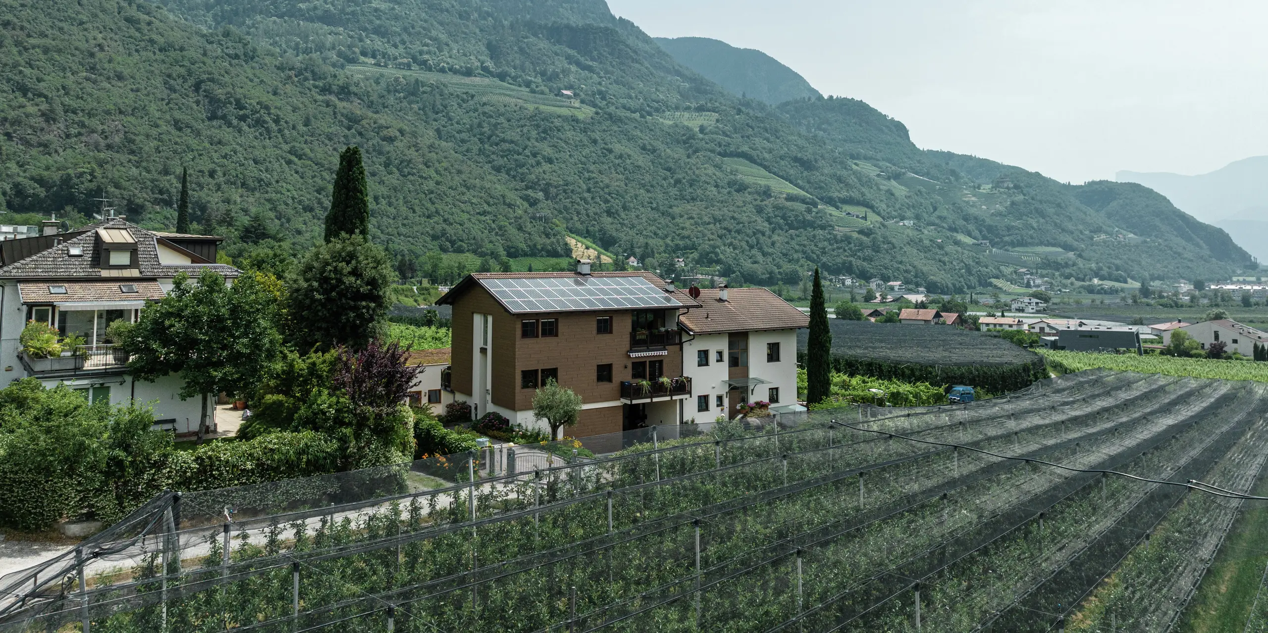 Vue aérienne des appartements de vacances Prechtlhof à Merano, dans le Tyrol du Sud, entourés de vergers et de paysages de montagne. La façade modernisée avec des Sidings PREFA en noyer foncé offre un aspect bois naturel en aluminium durable, robuste et nécessitant peu d'entretien. L'installation photovoltaïque sur le toit contribue à un approvisionnement énergétique durable.