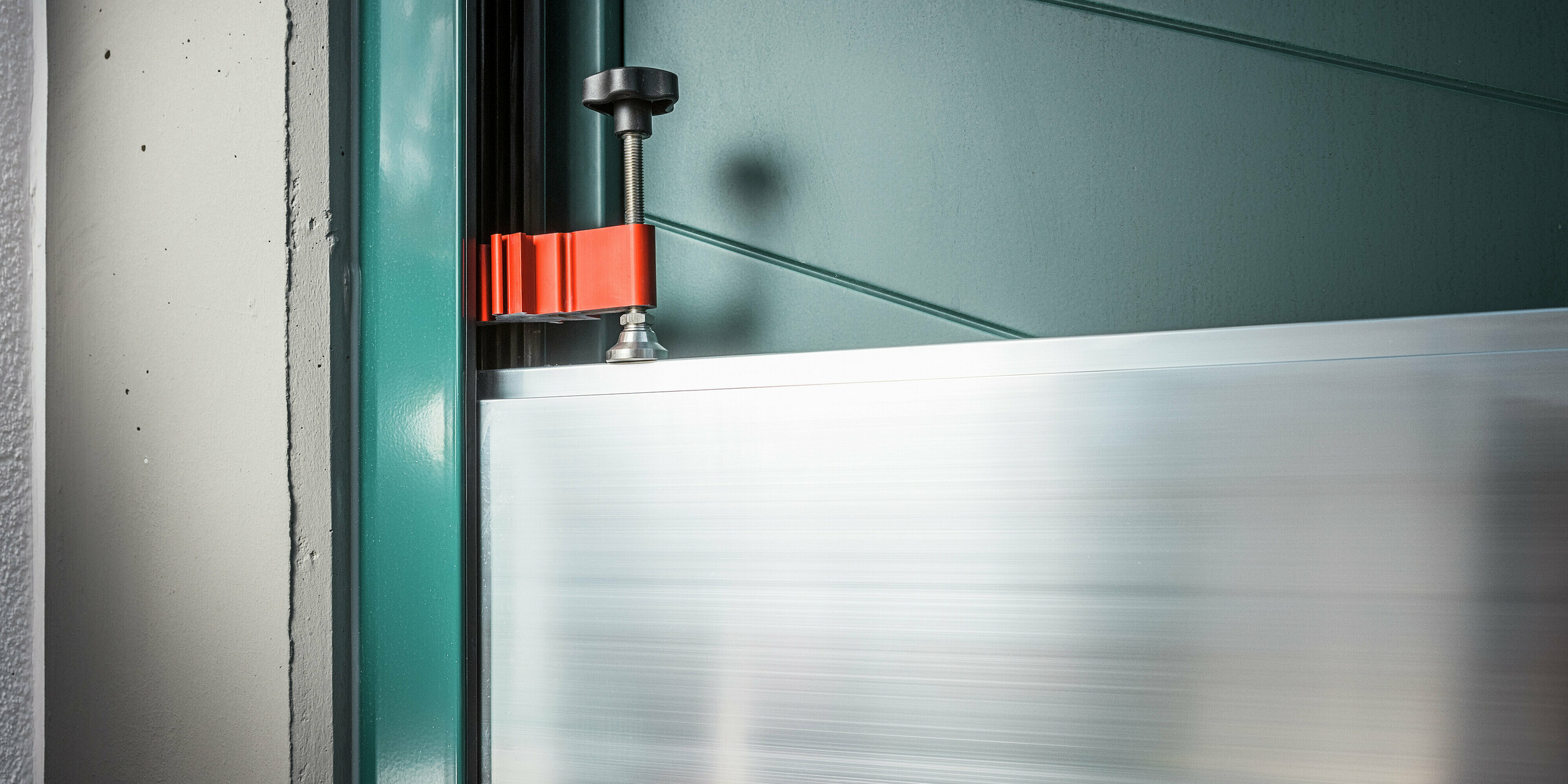 Gros plan sur le dispositif de serrage du système de protection contre les crues PREFA installé dans l'embrasure d'une maison individuelle à Schwaz. L'image montre la fixation précise des batardeaux en aluminium dans les profilés des parois latérales à l'aide d'une pièce se serrage rouge et d'un serre-joint noir. La qualité de fabrication et la simplicité d'utilisation soulignent l'aptitude du système à un usage quotidien : idéal pour une protection rapide contre les inondations aux entrées des maisons.