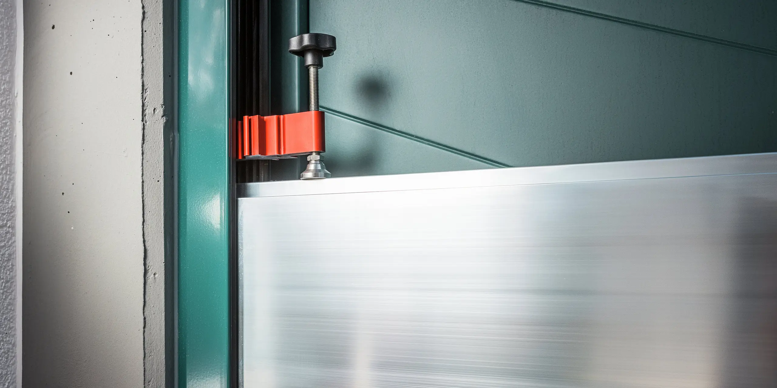 Gros plan sur le dispositif de serrage du système de protection contre les crues PREFA installé dans l'embrasure d'une maison individuelle à Schwaz. L'image montre la fixation précise des batardeaux en aluminium dans les profilés des parois latérales à l'aide d'une pièce se serrage rouge et d'un serre-joint noir. La qualité de fabrication et la simplicité d'utilisation soulignent l'aptitude du système à un usage quotidien : idéal pour une protection rapide contre les inondations aux entrées des maisons.