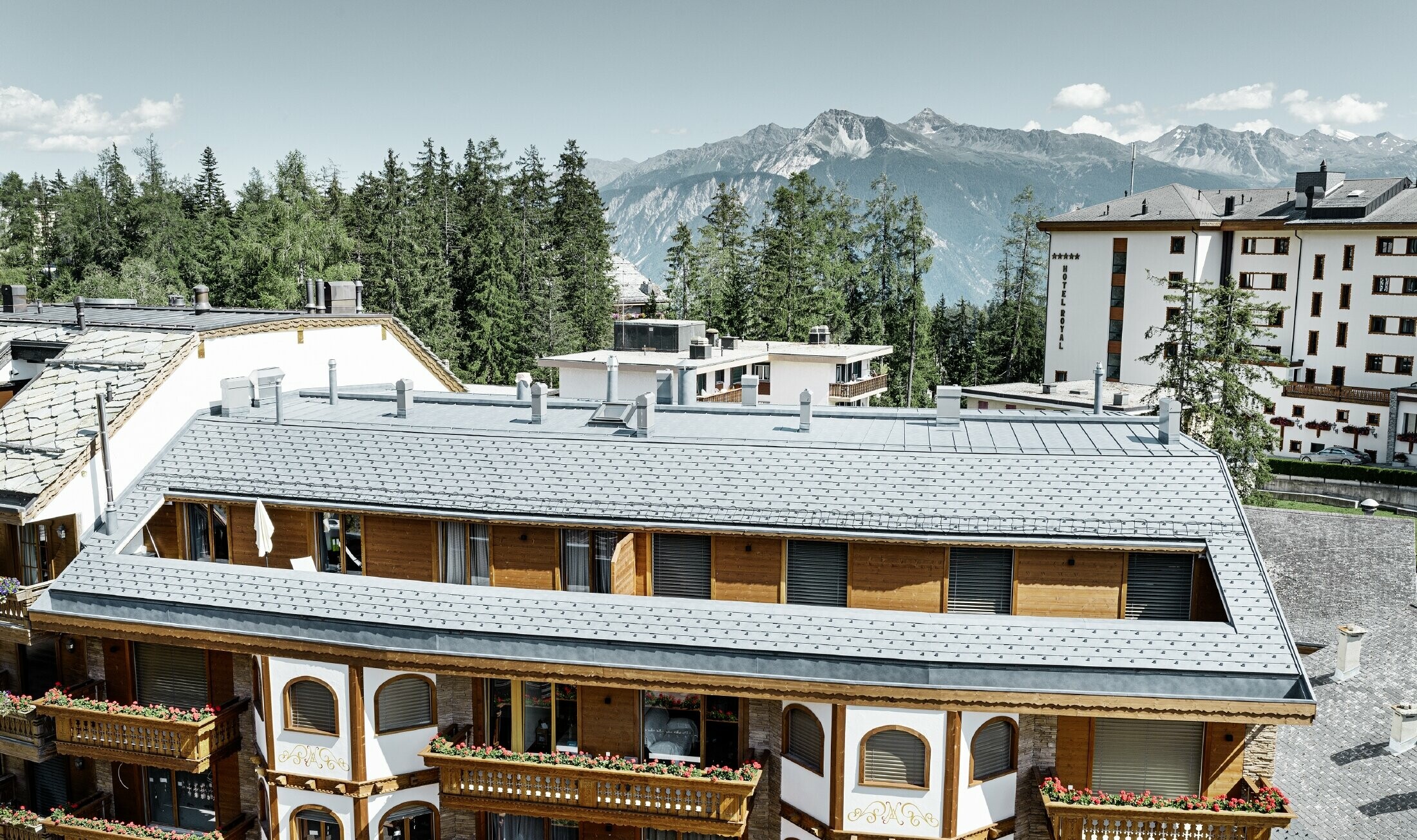 Immeuble d’habitation de Crans-Montana (Suisse) avec les Alpes en arrière-plan — Façade intégrant de nombreux éléments en bois et dont la toiture a été réalisée avec des bardeaux en aluminium PREFA de couleur gris pierre