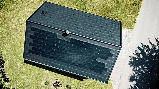 [Translate to Français:] Die spektakuläre Drohnenaufnahme zeigt das PREFA Solardach von Haus Immi aus der Vogelperspektive. Schwarze Solardachplatten gehen nahezu unsichtbar in die Dachplatten R.16 in P.10 Schwarz über und formen eine durchgängige Dachfläche. Das integrierte Aluminiumsystem vereint nachhaltige Solarstromerzeugung, hohe Widerstandsfähigkeit und klare Architektur ohne optisch aufgesetzte Photovoltaikanlage im alpinen Raum.