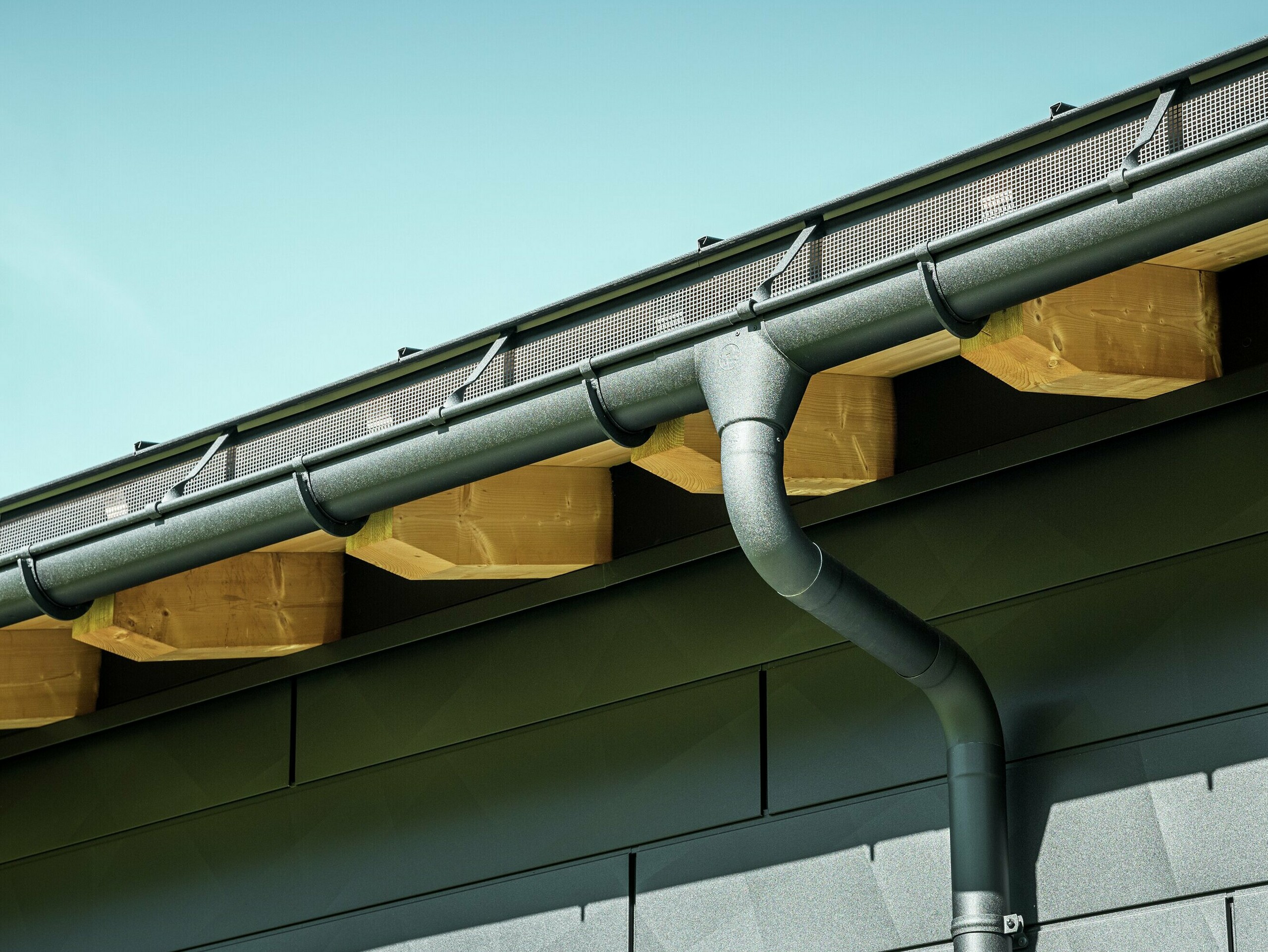 Gouttière demi-ronde PREFA avec naissance et étrier en P.10 noir Gros plan de la façade d'une maison familiale moderne à Ascharina, en Suisse. Les côtés du bâtiment ont été revêtus des remarquables panneaux de façade PREFA Siding.X en P.10 noir. Le système de gouttière PREFA avec gouttière suspendue et tuyau de descente est de couleur assortie.