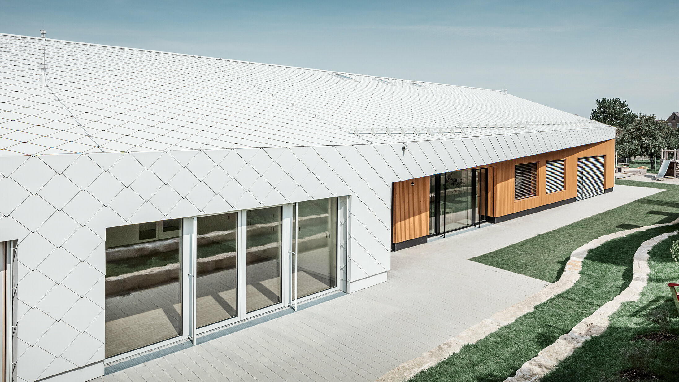 Jardin d’enfants revêtu du losange de toiture et de façade 44 de PREFA, en blanc Prefa ; la surface du toit et la façade ne font qu'un ; grandes surfaces vitrées